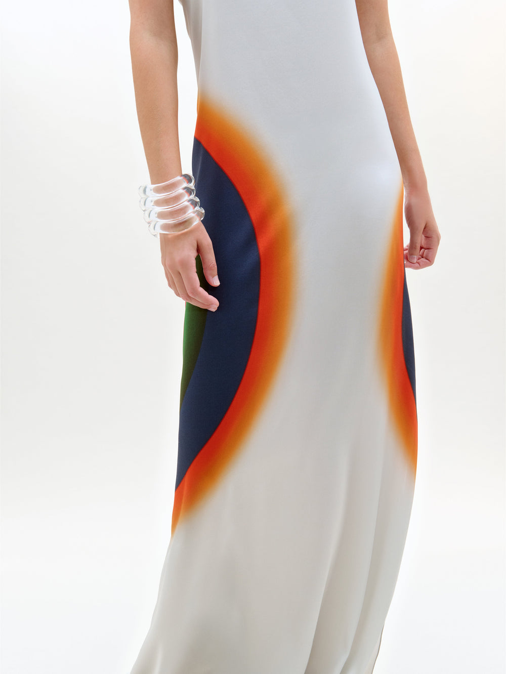 Una persona lleva el vestido Bahiana, un maxi de seda blanca sin mangas con atrevidos motivos circulares en naranja, rojo, azul marino y verde. Fabricado en Portugal, se combina con brazaletes transparentes sobre un fondo blanco liso.