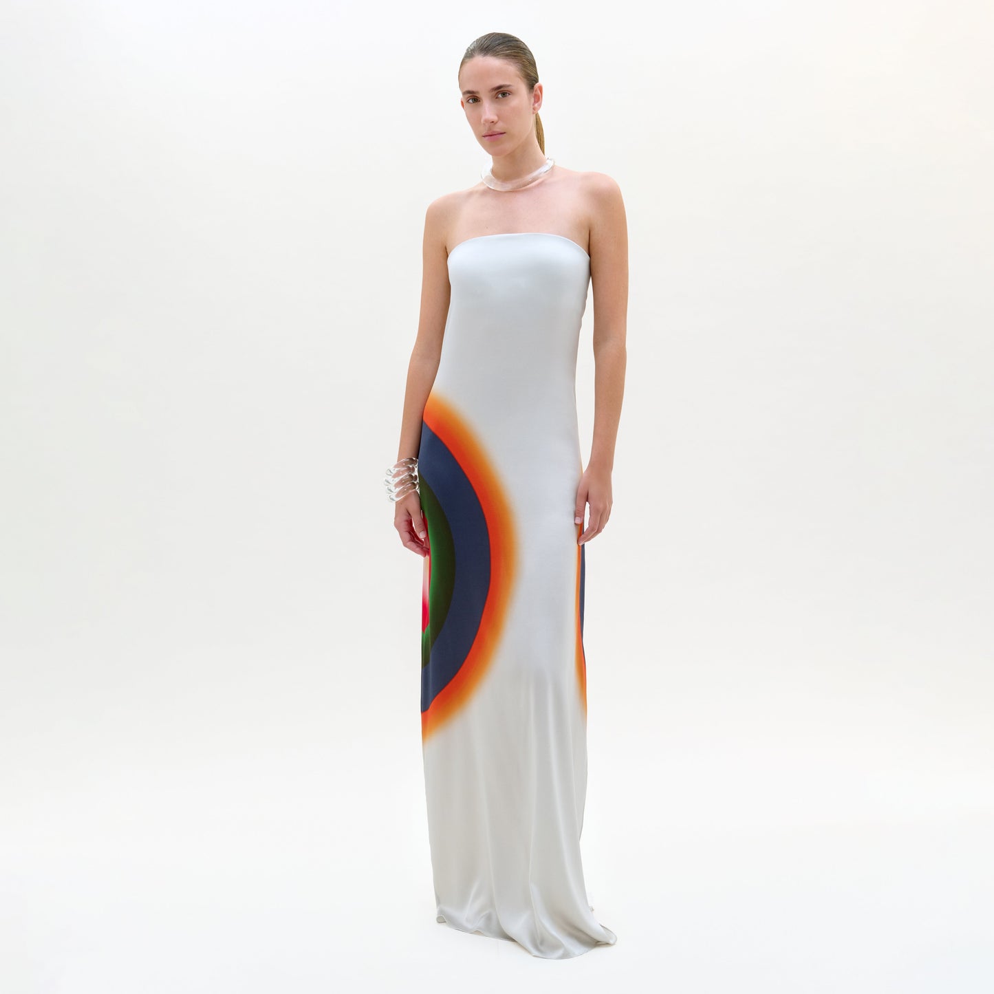 Una mujer, sobre un fondo liso, lleva el vestido Bahiana, un maxi de seda hecho en Portugal con un colorido estampado circular cerca del dobladillo. Lleva el pelo recogido y accesorios como pulseras y un collar.
