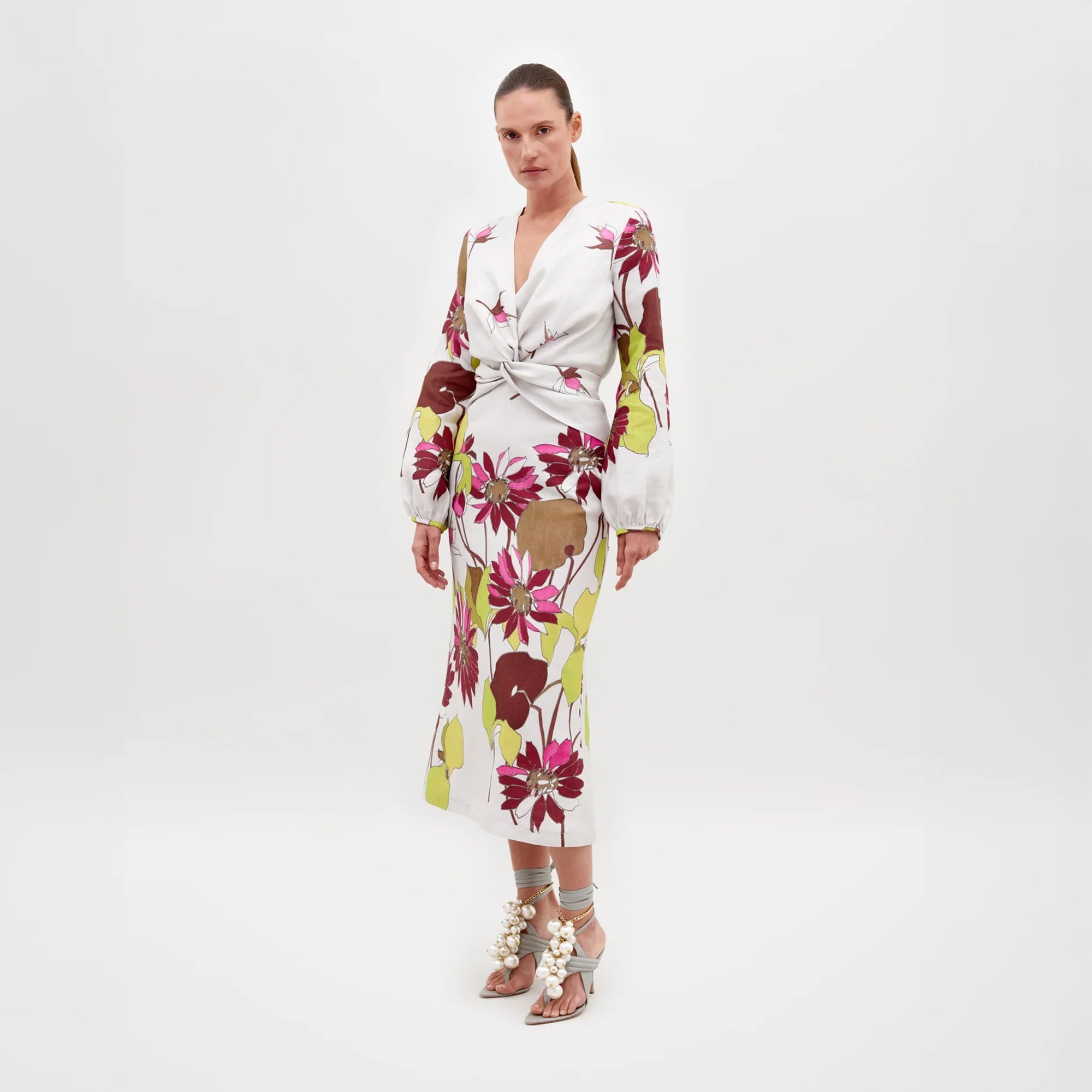 Una mujer posa sobre un fondo blanco liso con el Vestido Battia, un elegante vestido midi hecho en Colombia con llamativos estampados florales en rojo, rosa y verde, mangas largas abullonadas y detalles recortados, combinado con sandalias de tacón adornadas en gris.