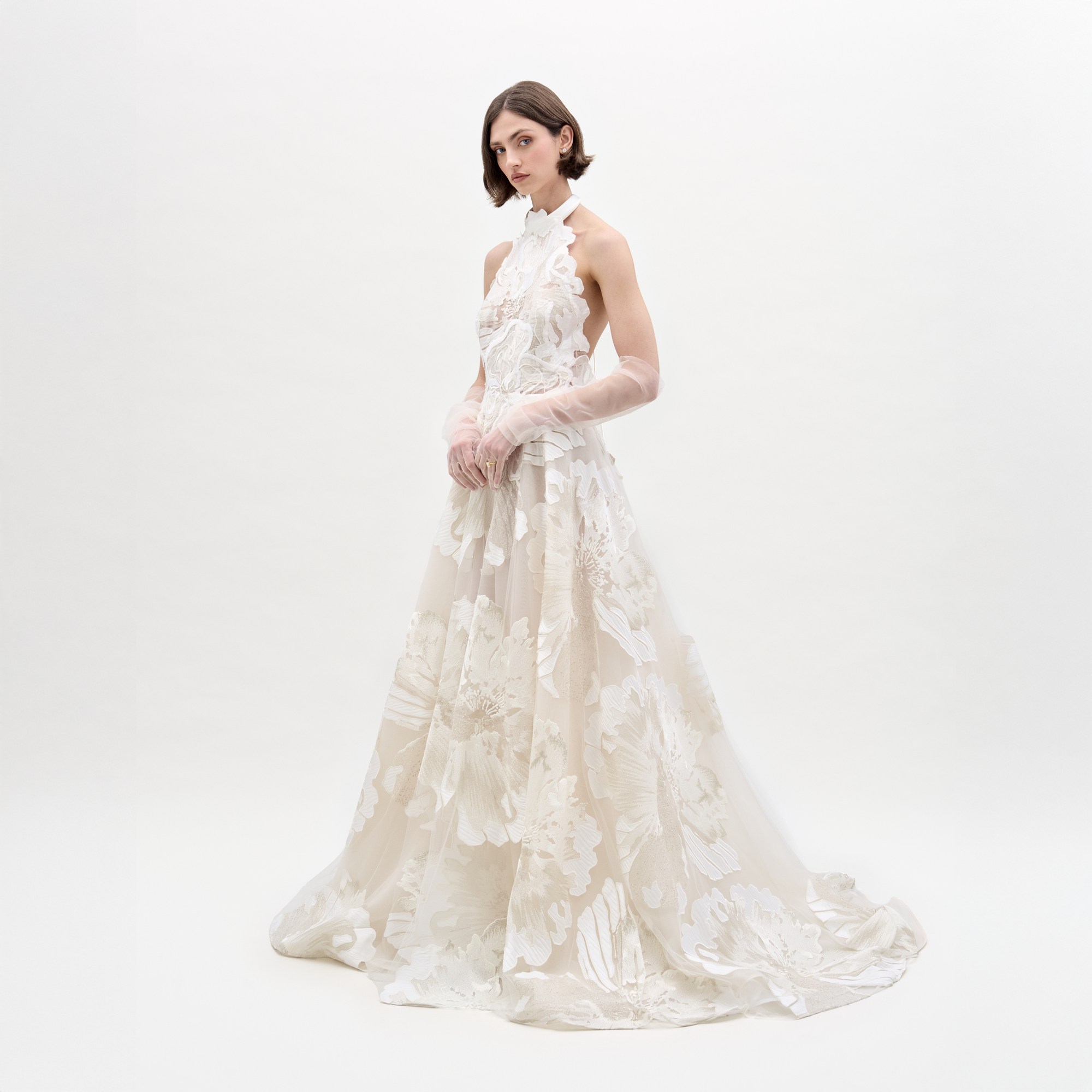 Una mujer con cabello castaño corto se encuentra sobre un fondo blanco liso con el Vestido Becca, un vestido largo hasta el suelo con cuello alto y encaje floral translúcido, combinado con guantes transparentes hasta el codo.