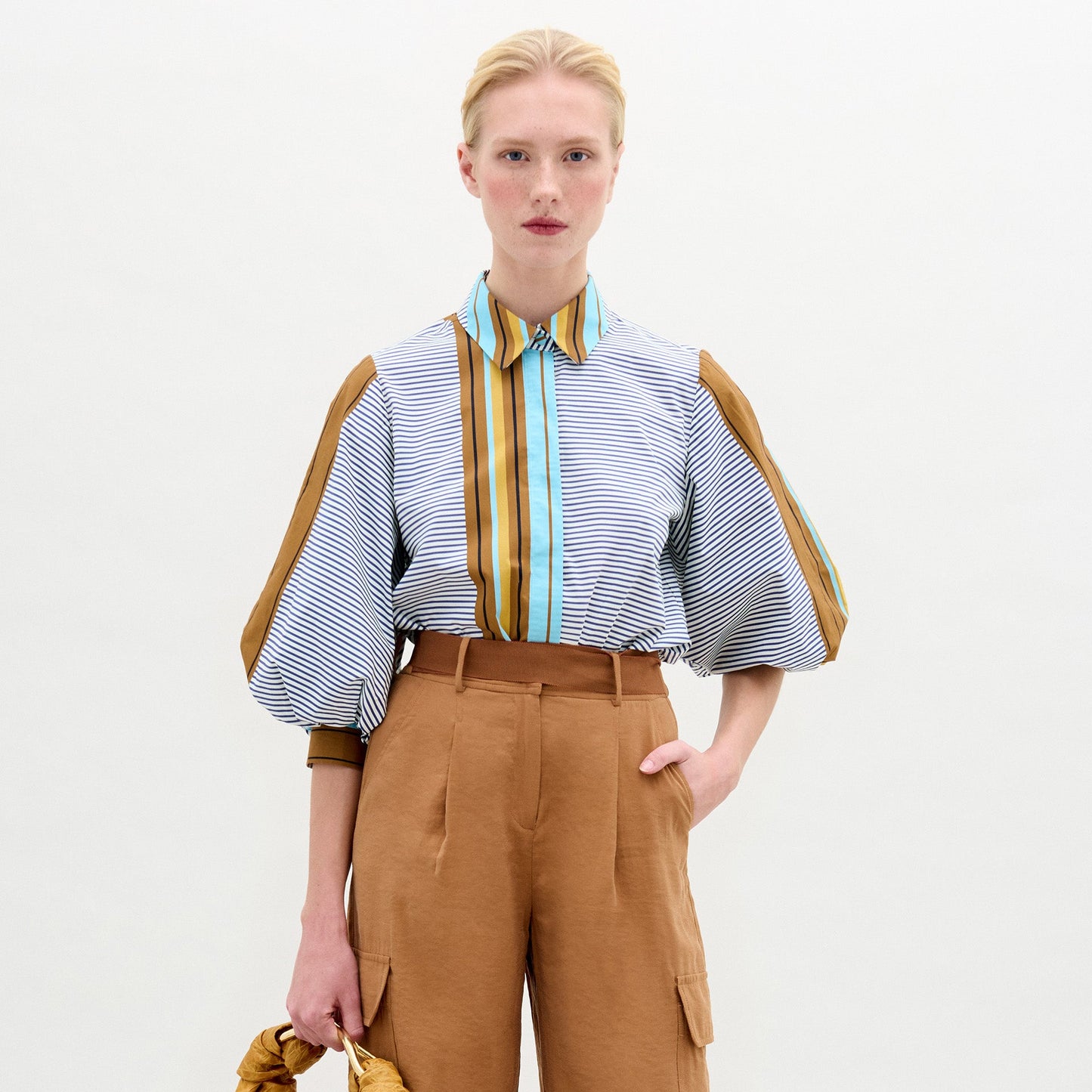 Una persona de piel clara y pelo rubio lleva la blusa Belkis, con mangas abullonadas y detalles verticales marrones, combinada con unos pantalones de cintura alta de color canela. Sobre fondo blanco, sostiene un objeto marrón en la mano izquierda.