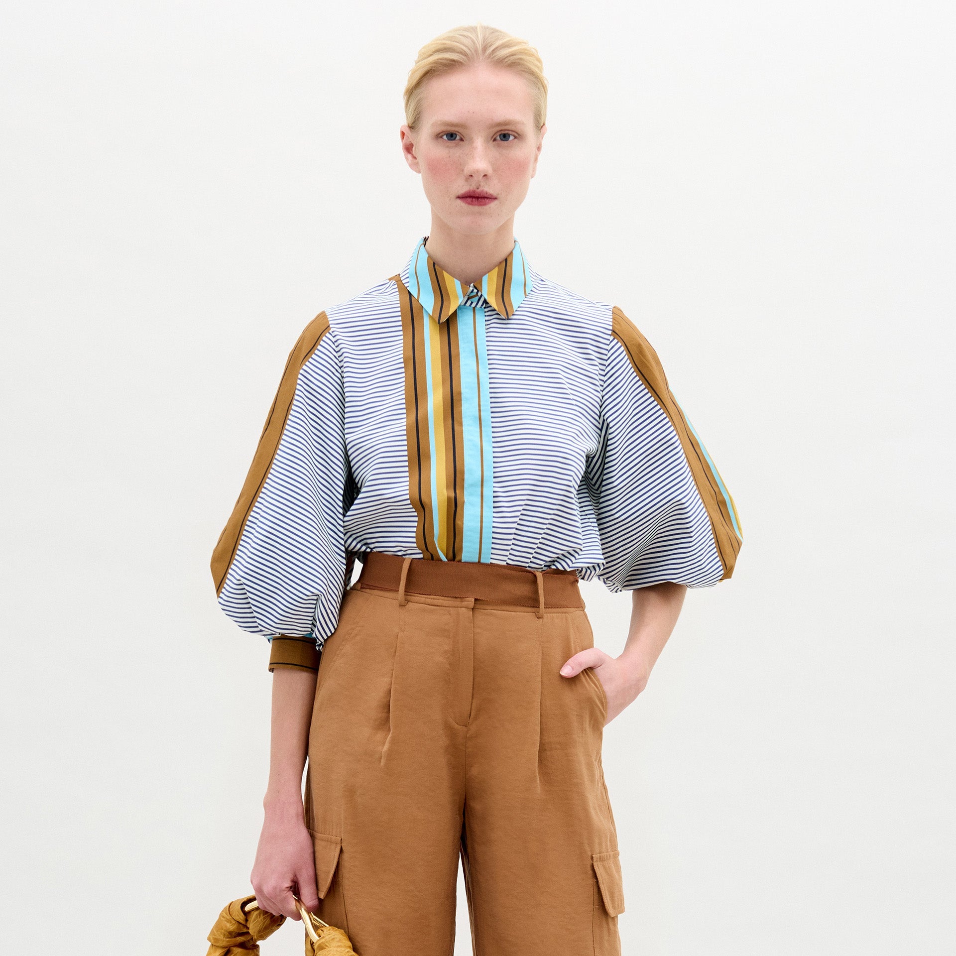 Una persona de piel clara y pelo rubio lleva la blusa Belkis, con mangas abullonadas y detalles verticales marrones, combinada con unos pantalones de cintura alta de color canela. Sobre fondo blanco, sostiene un objeto marrón en la mano izquierda.