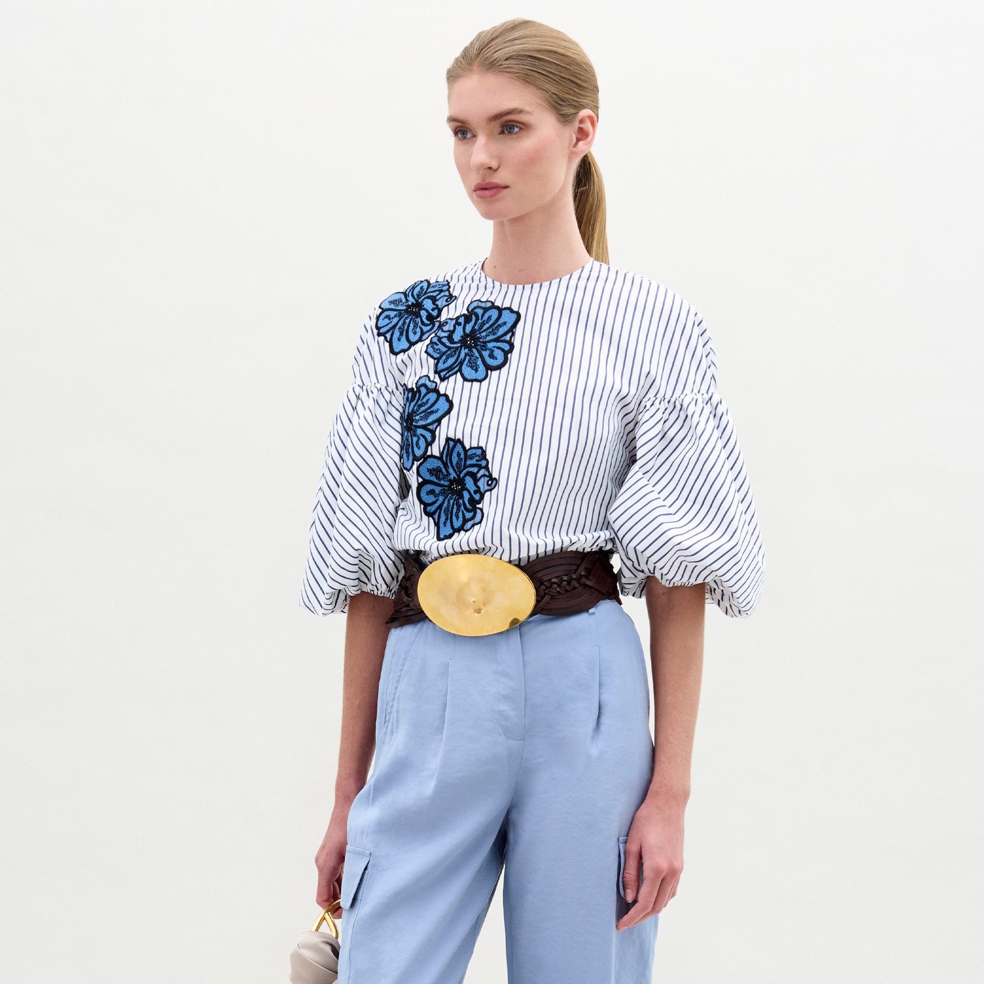 Una persona, de pie sobre un fondo liso, lleva la blusa Belle con bordados florales azules, pantalones de cintura alta azul claro, un cinturón marrón ancho con una gran hebilla redonda y un pequeño bolso beige en la mano.