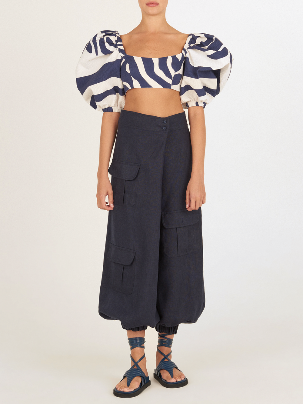 Una persona está de pie mirando al frente con un crop top de cebra de mangas abullonadas azul y blanco, joggers de lino azul marino con bolsillos y sandalias de tiras azules. Su cabeza aparece recortada en la imagen.