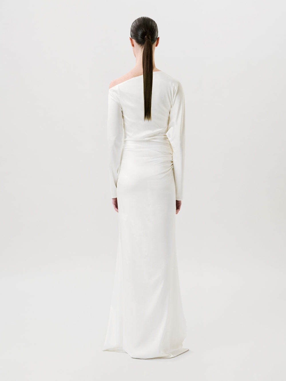 A person with long dark hair in a low ponytail wears the floor-length, long-sleeved Bahir Dress in white silk and stands facing away against a plain light background, showcasing timeless elegance in this ready-to-wear piece made in the USA.
