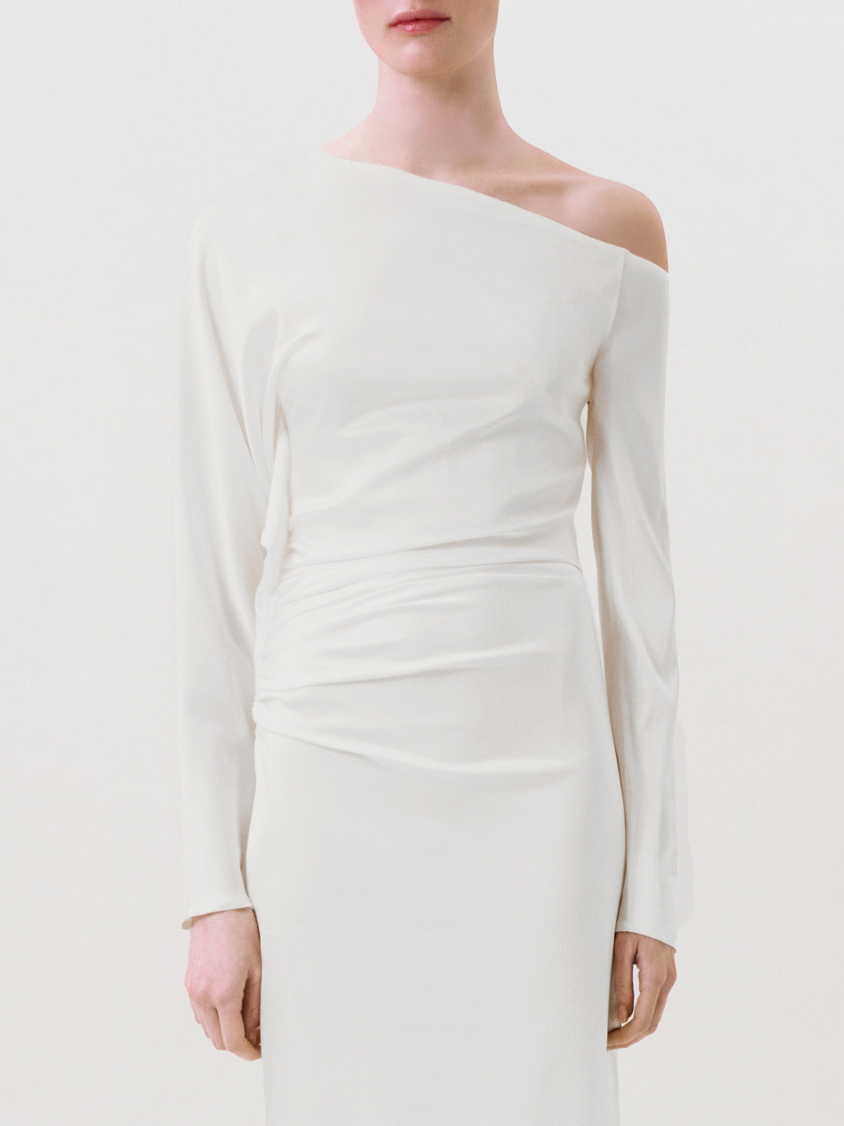 A person models the Bahir Dress—a minimalist white silk, one-shoulder long-sleeve design—standing against a plain background. The ready-to-wear piece is displayed in an image cropped from mid-face to mid-thigh.