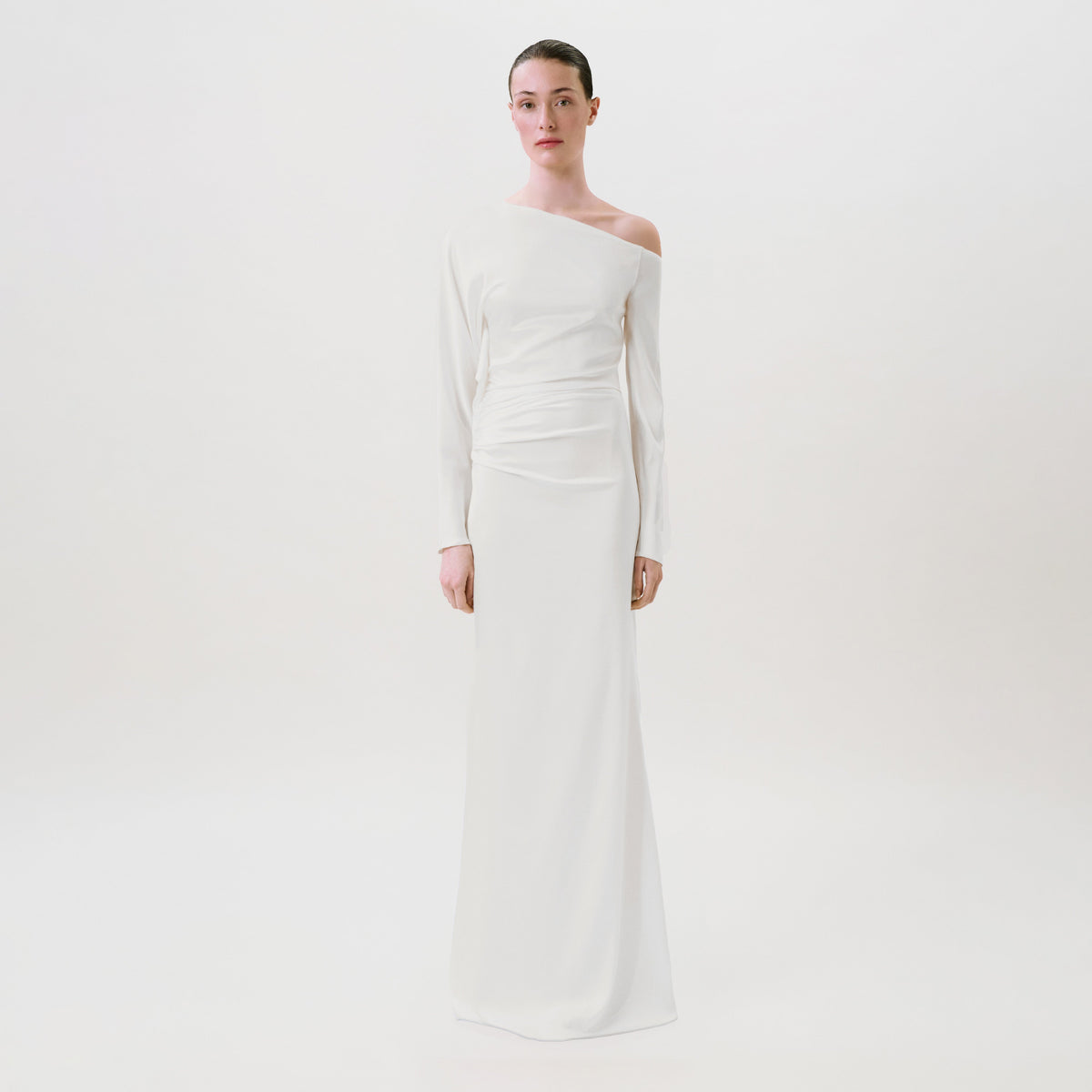 A woman stands against a plain white background wearing the Bahir Dress, a long, white, one-shoulder gown crafted from luxurious silk. This elegant ready-to-wear piece features long sleeves and a draped waist detail, expertly made in the USA for effortless sophistication.