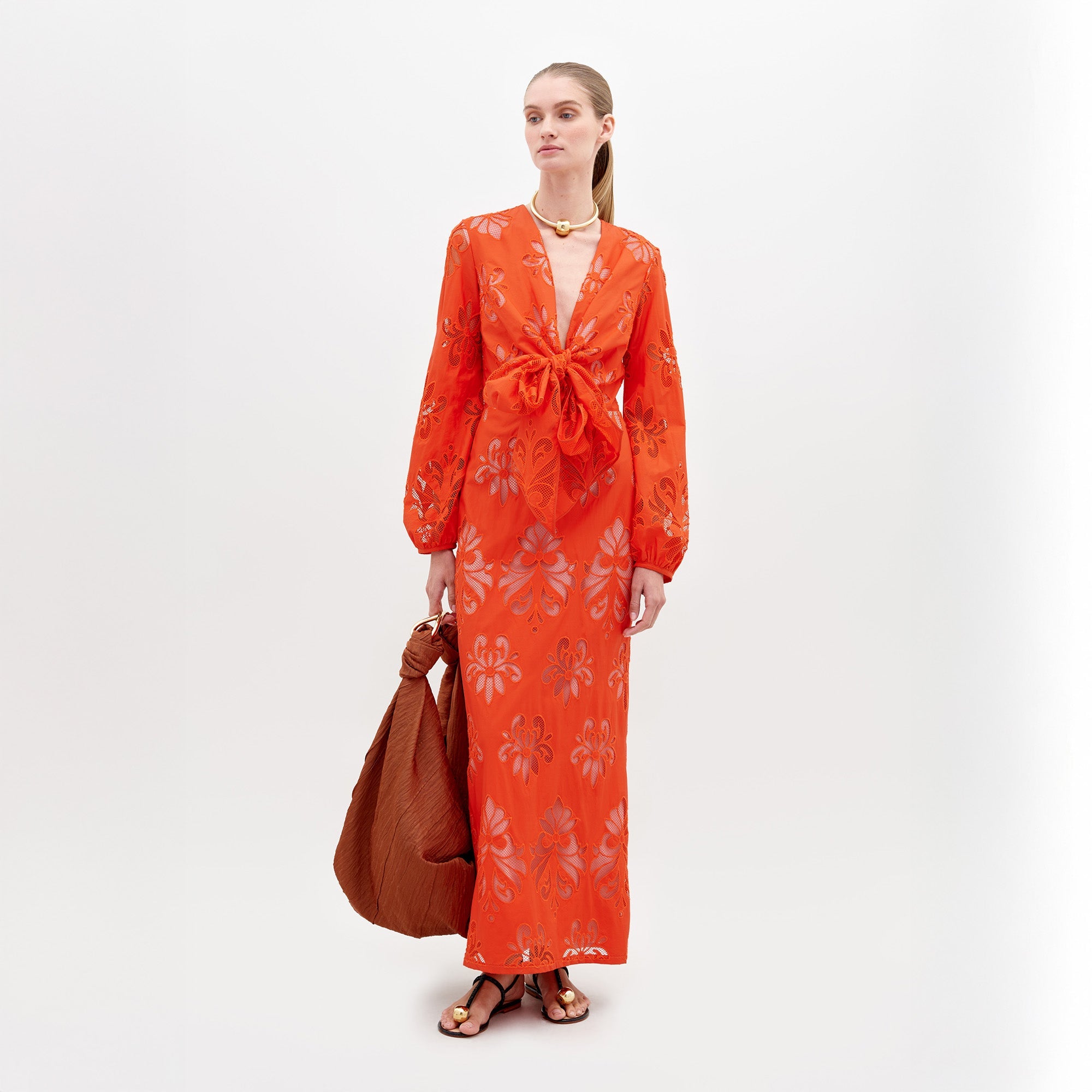 Una mujer se encuentra de pie sobre un fondo blanco liso con el Vestido Battia, un vestido midi naranja brillante con estampados florales, mangas largas y un gran lazo delantero. Lleva un gran bolso marrón, sandalias planas y un collar dorado.