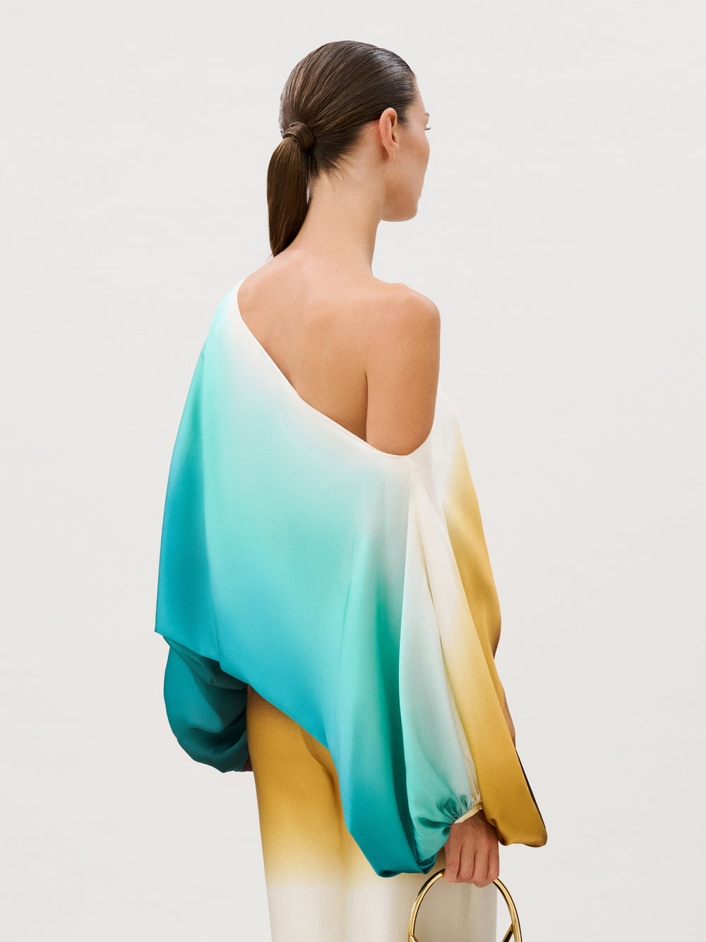 The Bellagio Blouse is an Italian silk one-shoulder top with a flowing, ombre design that transitions from teal to white to yellow, worn with matching wide-leg trousers.