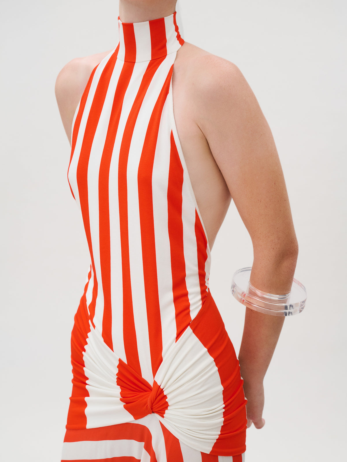 The Belona Dress is a sleeveless halter dress in red and white vertical striped Italian crepe de viscose, featuring a gathered knot detail at the waist and a clear circular bracelet on one arm.