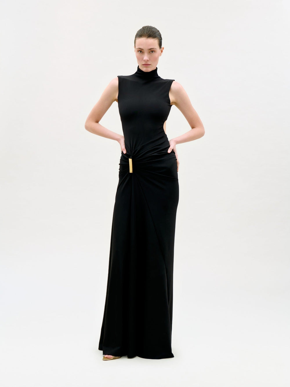 A woman poses against a plain white background, wearing the Bogna Dress—a sleeveless, floor-length black maxi dress with a high neckline and gold waist accent—looking confidently at the camera with her hands on her hips.