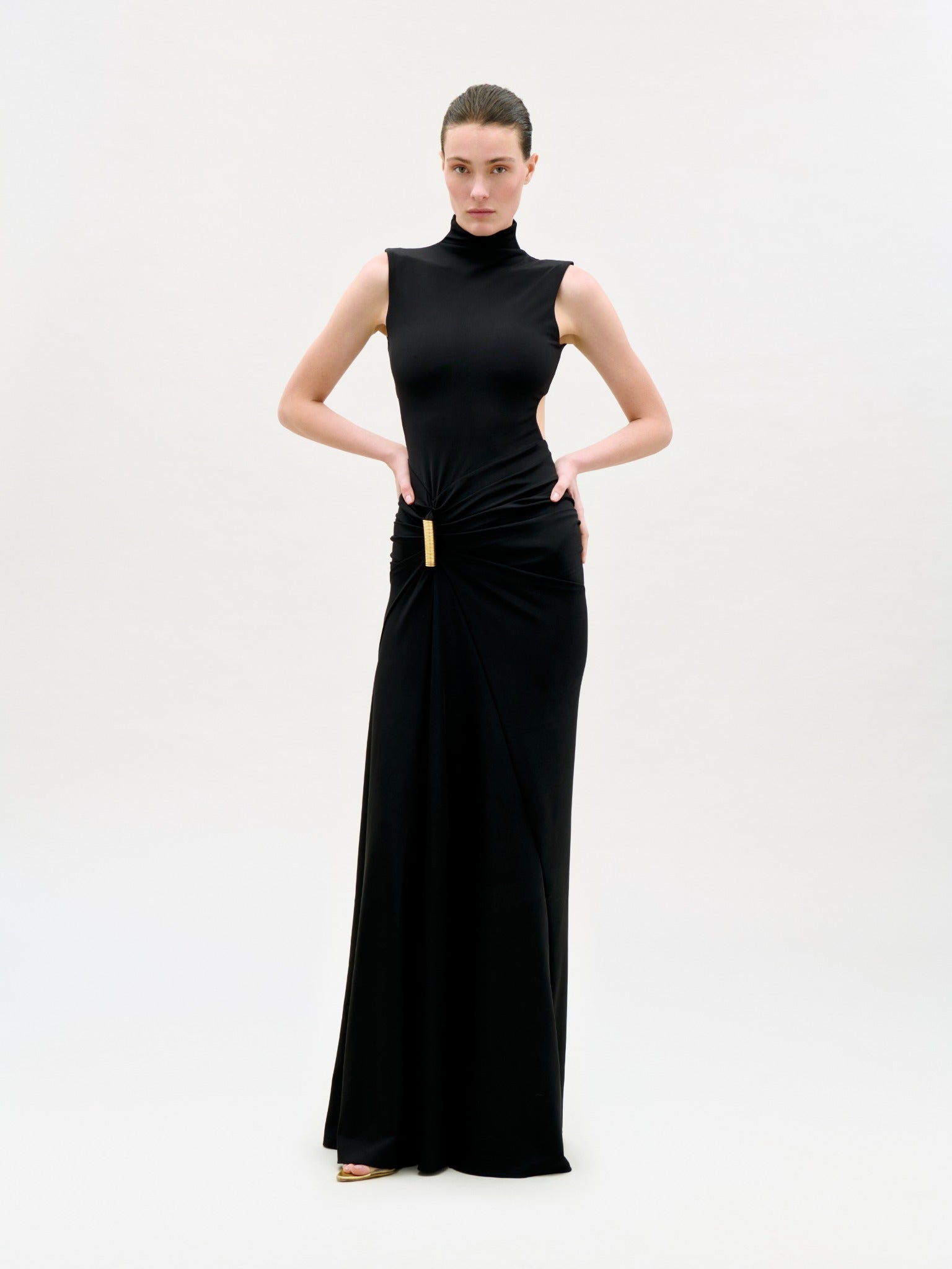 A woman poses against a plain white background, wearing the Bogna Dress—a sleeveless, floor-length black maxi dress with a high neckline and gold waist accent—looking confidently at the camera with her hands on her hips.