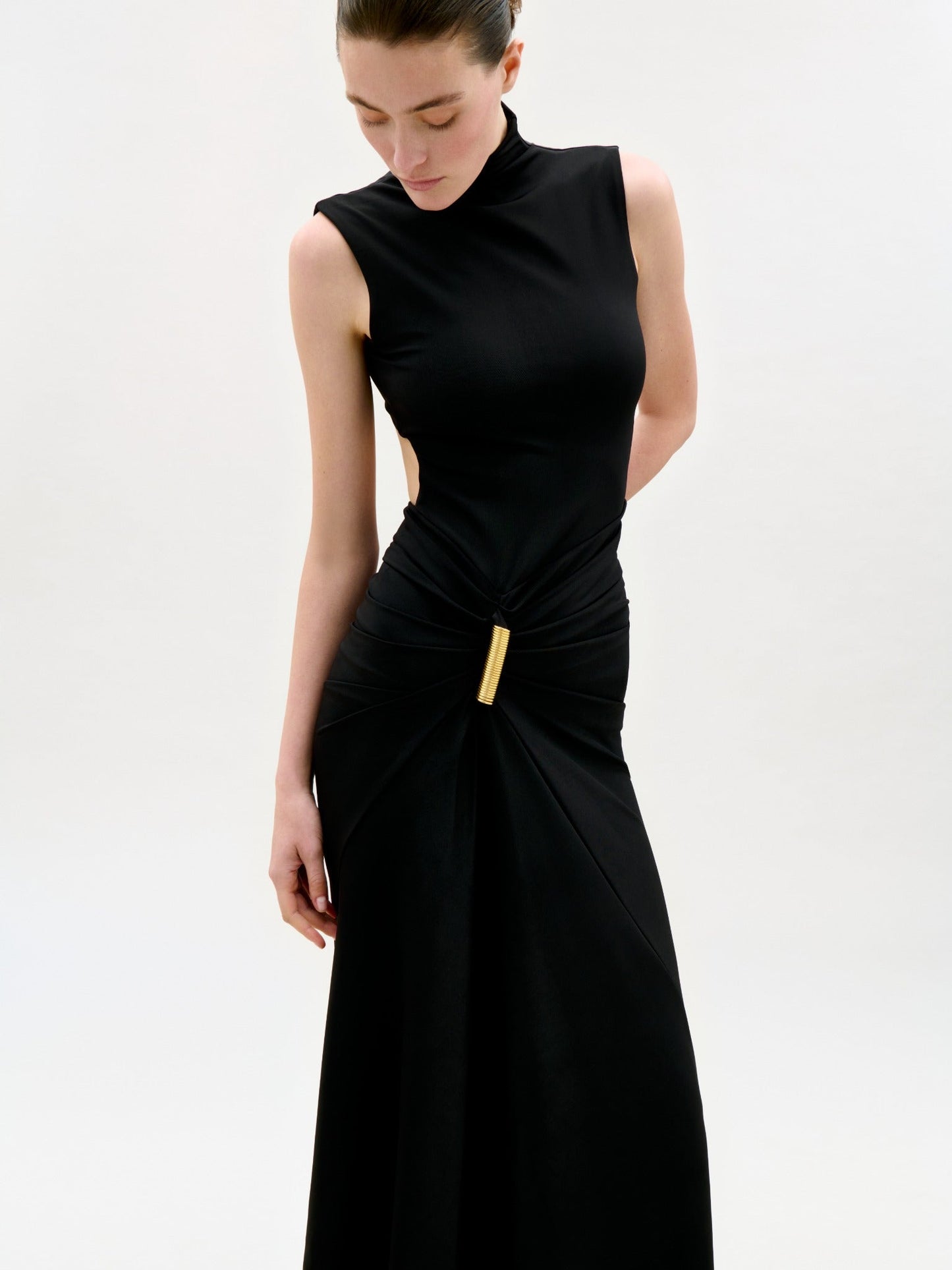 A woman stands against a plain light background, looking down with her hair pulled back, wearing the Bogna Dress—a sleeveless black maxi with a high neckline, gathered waist, gold clasp accent, and asymmetric skirt.