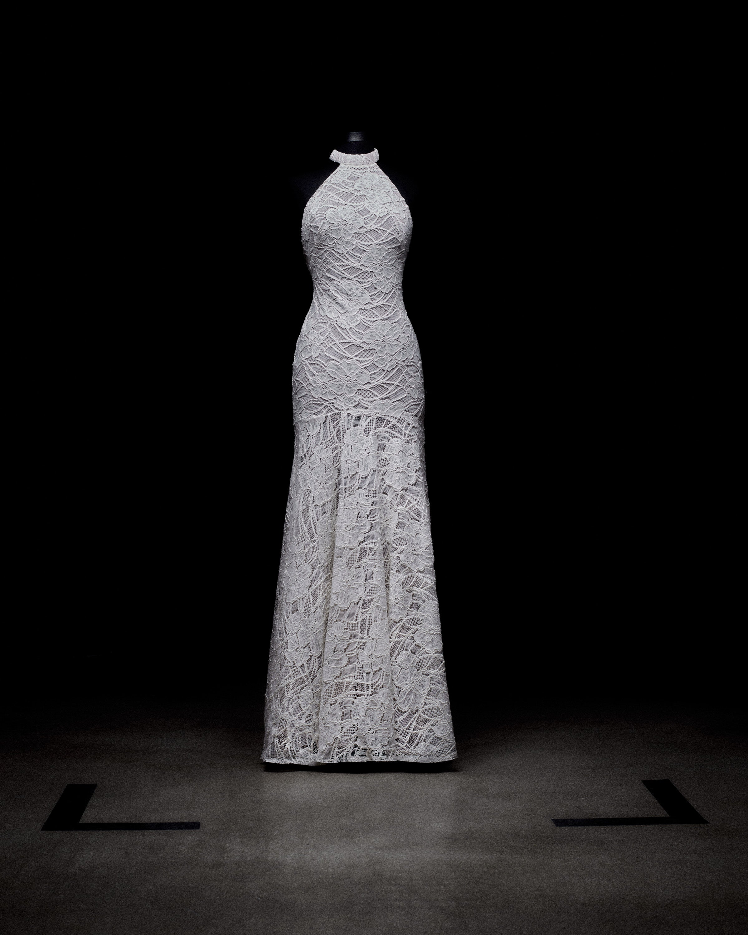 Un vestido de noche de encaje blanco se exhibe sobre un maniquí negro y se coloca sobre un fondo oscuro. El vestido presenta un escote alto y una silueta ajustada que se extiende hasta una falda larga y elegante. El foco resalta sus intrincados detalles de encaje.