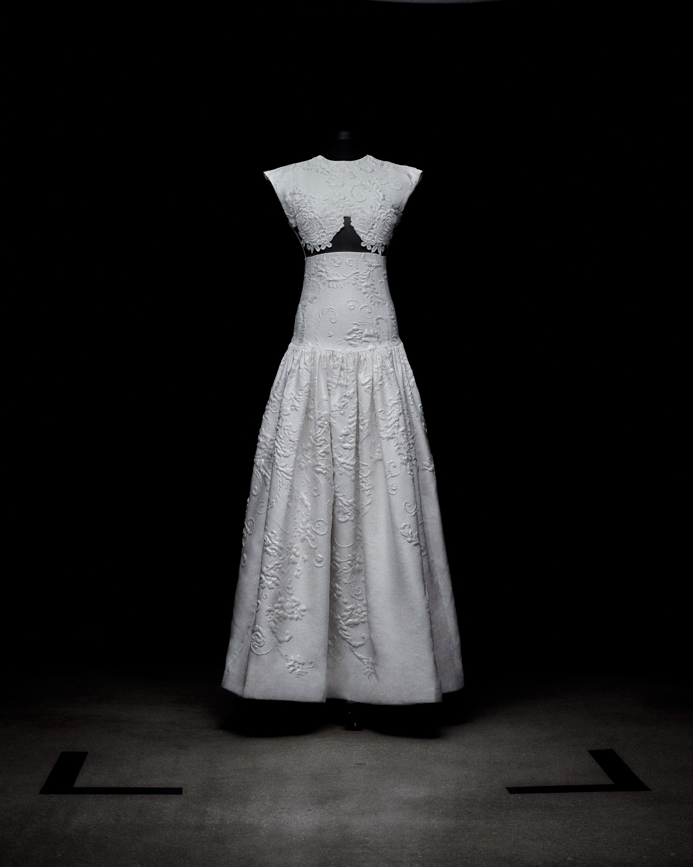 Un vestido largo y blanco exhibido sobre un maniquí contra un fondo oscuro. El vestido presenta un bordado intrincado, un corpiño ajustado con escote alto y un detalle de corte debajo del busto. La falda es larga y elegantemente drapeada.