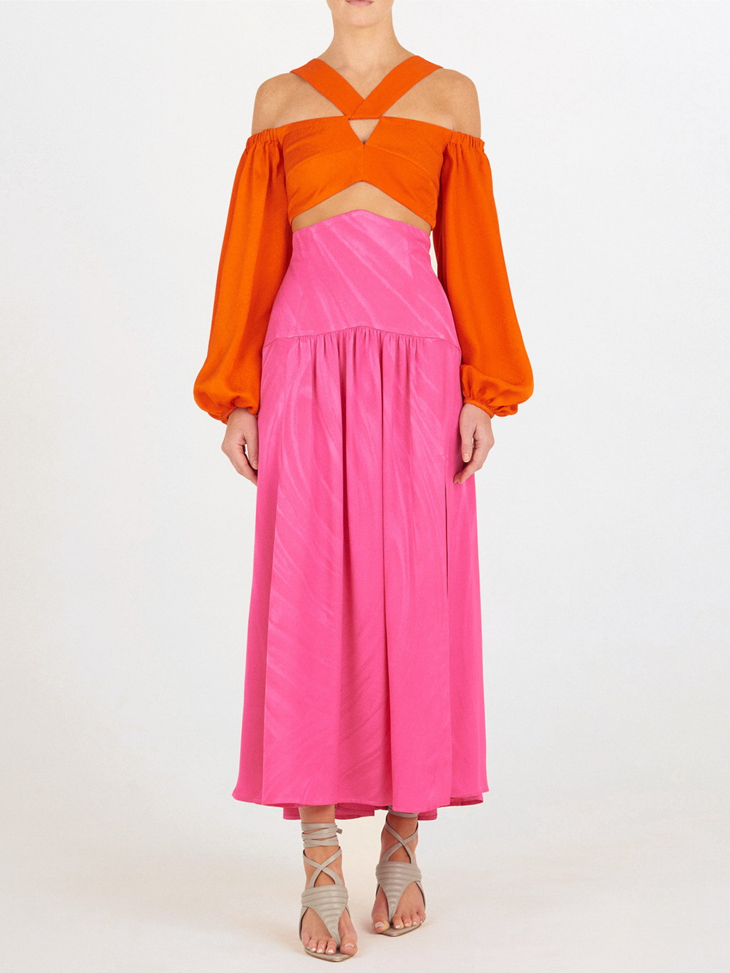 Una persona, sobre un fondo liso, lleva la Falda Callie Fuchsia, una falda de jacquard de alta costura hasta el suelo, combinada con un top naranja de hombros recortados, manga larga y tiras cruzadas, y sandalias de tiras beige.