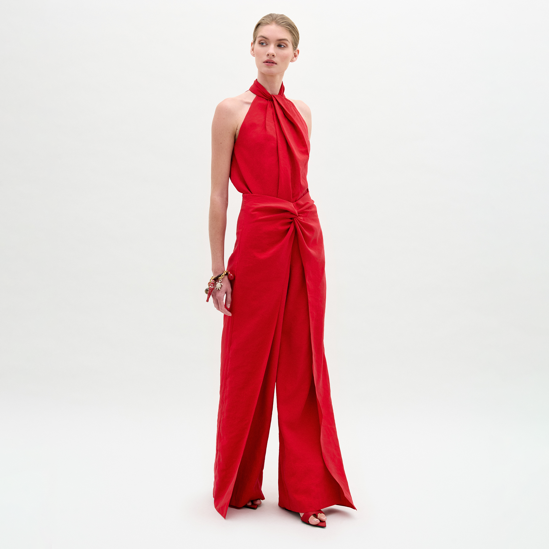 Una persona, sobre un fondo blanco liso, lleva el Pantalón Canturipe, un mono rojo sin mangas, con cuello halter, pantalón ancho de tiro alto y detalle de vuelta en la cintura, combinado con sandalias rojas y una pulsera.
