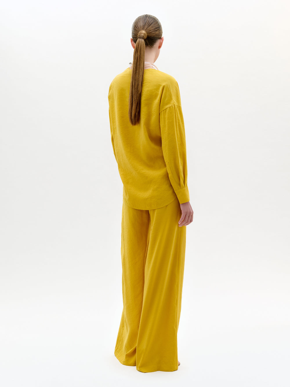 Una persona con el pelo largo recogido en una coleta mira hacia otro lado, con un top amarillo holgado de manga larga y unos pantalones Canturipe Pant, de pata ancha y tiro alto, fabricados en Colombia, sobre un fondo blanco liso.