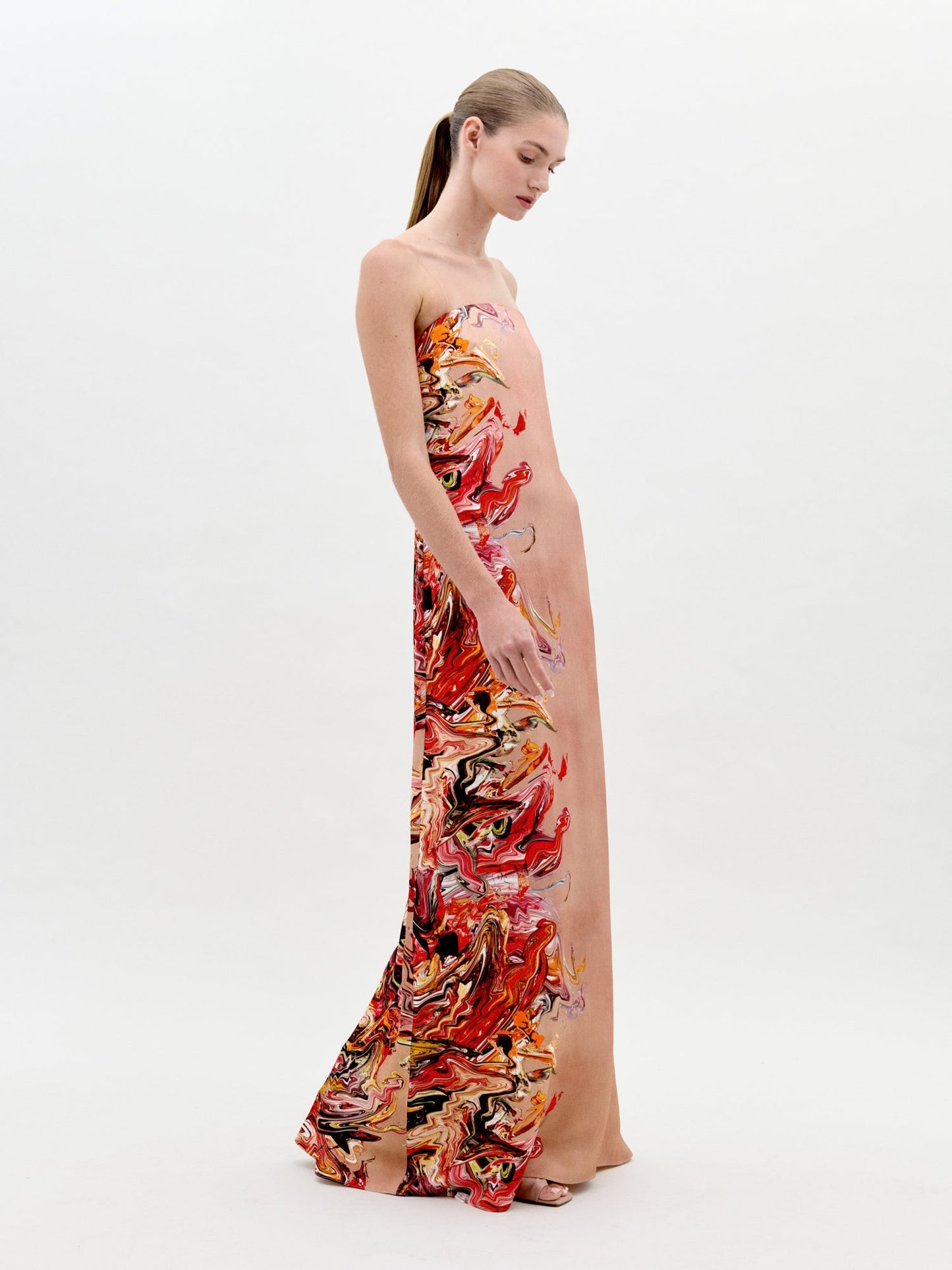 A woman stands in a long, strapless Adalgisa Dress Abstract Coral with red, orange, and yellow floral accents on the side against a plain white background. This stunning piece is available for pre-order now with an estimated ship-by date of February 1st, 2025.