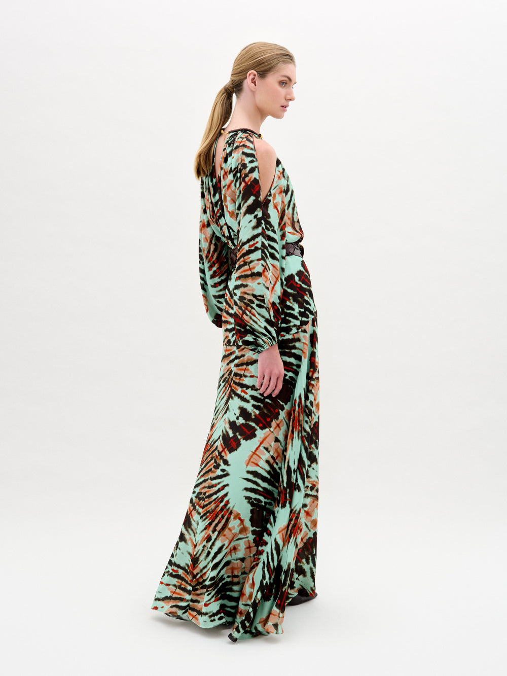 Una persona aparece de perfil con el vestido Carla, un maxi largo y vaporoso con estampado tie-dye en verde, negro y naranja. El vestido tiene mangas anchas, hombros recortados y elegantes detalles dorados. El fondo es blanco.