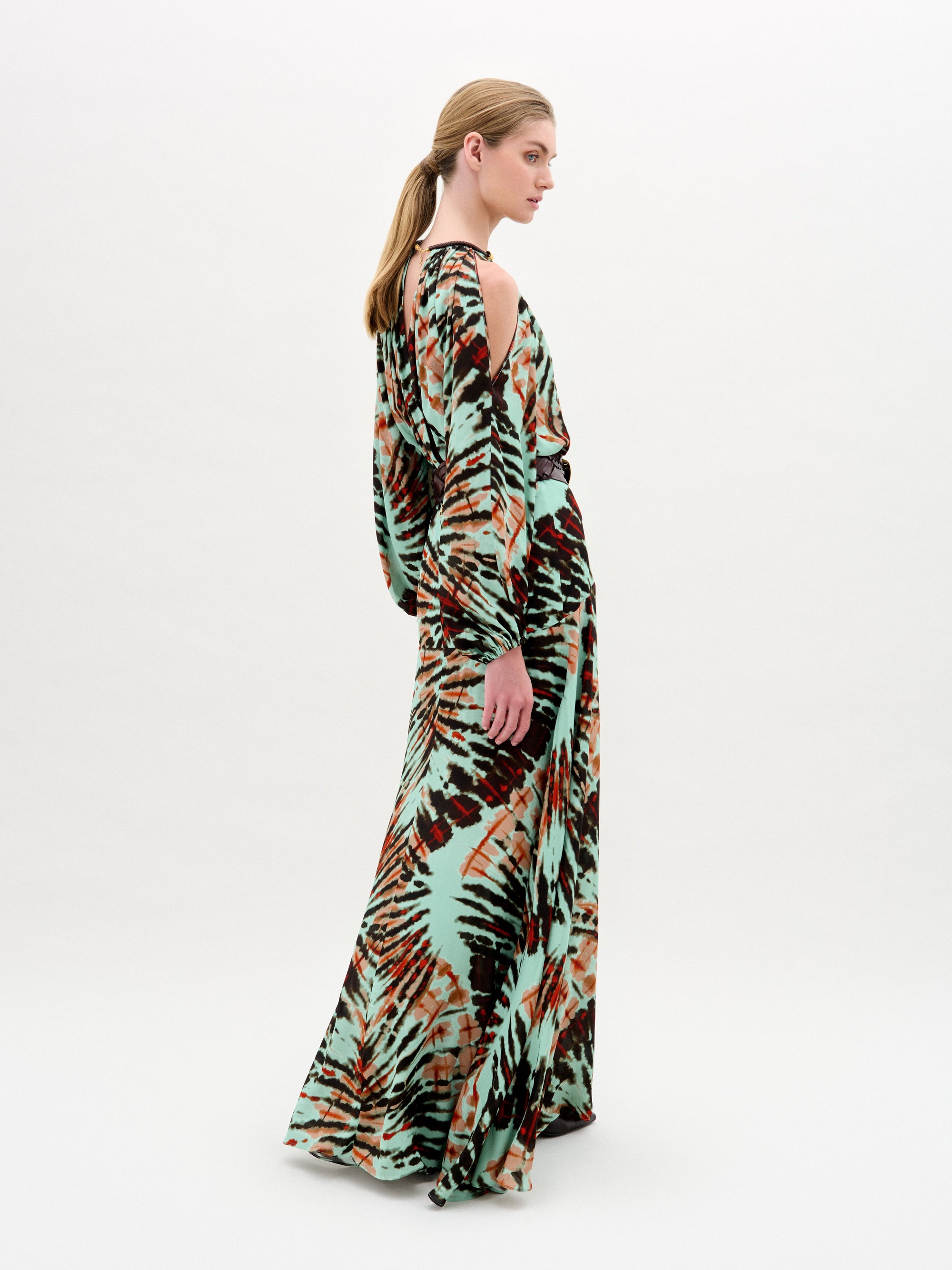Una persona aparece de perfil con el vestido Carla, un maxi largo y vaporoso con estampado tie-dye en verde, negro y naranja. El vestido tiene mangas anchas, hombros recortados y elegantes detalles dorados. El fondo es blanco.