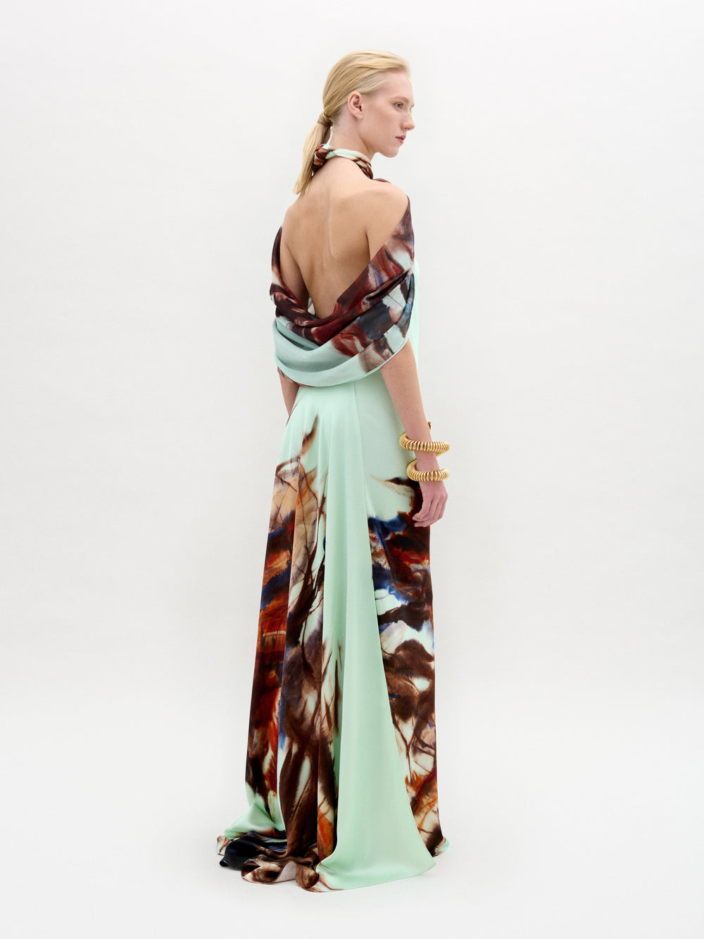 Una persona de perfil luce el vestido Carlina, un maxi largo con escote alto y espalda abierta, que muestra motivos abstractos en marrón, azul y crema sobre tejido italiano verde claro. Los brazaletes dorados completan el look sobre un fondo blanco.