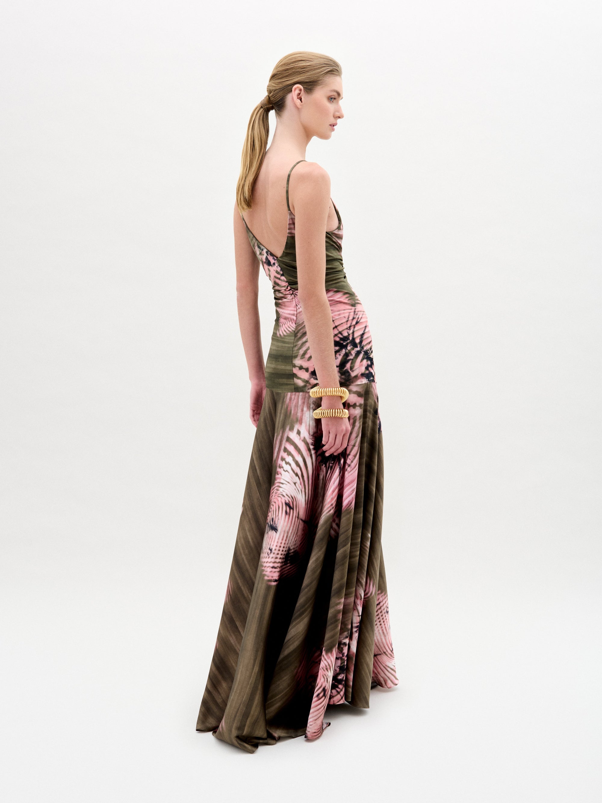 Una mujer con el pelo claro recogido en una coleta baja lleva el Carmin Dress, un maxi vestido sin mangas de tela italiana con estampado rosa y marrón. Está de lado, mirando al frente, y lleva pulseras en la mano izquierda sobre un fondo blanco.