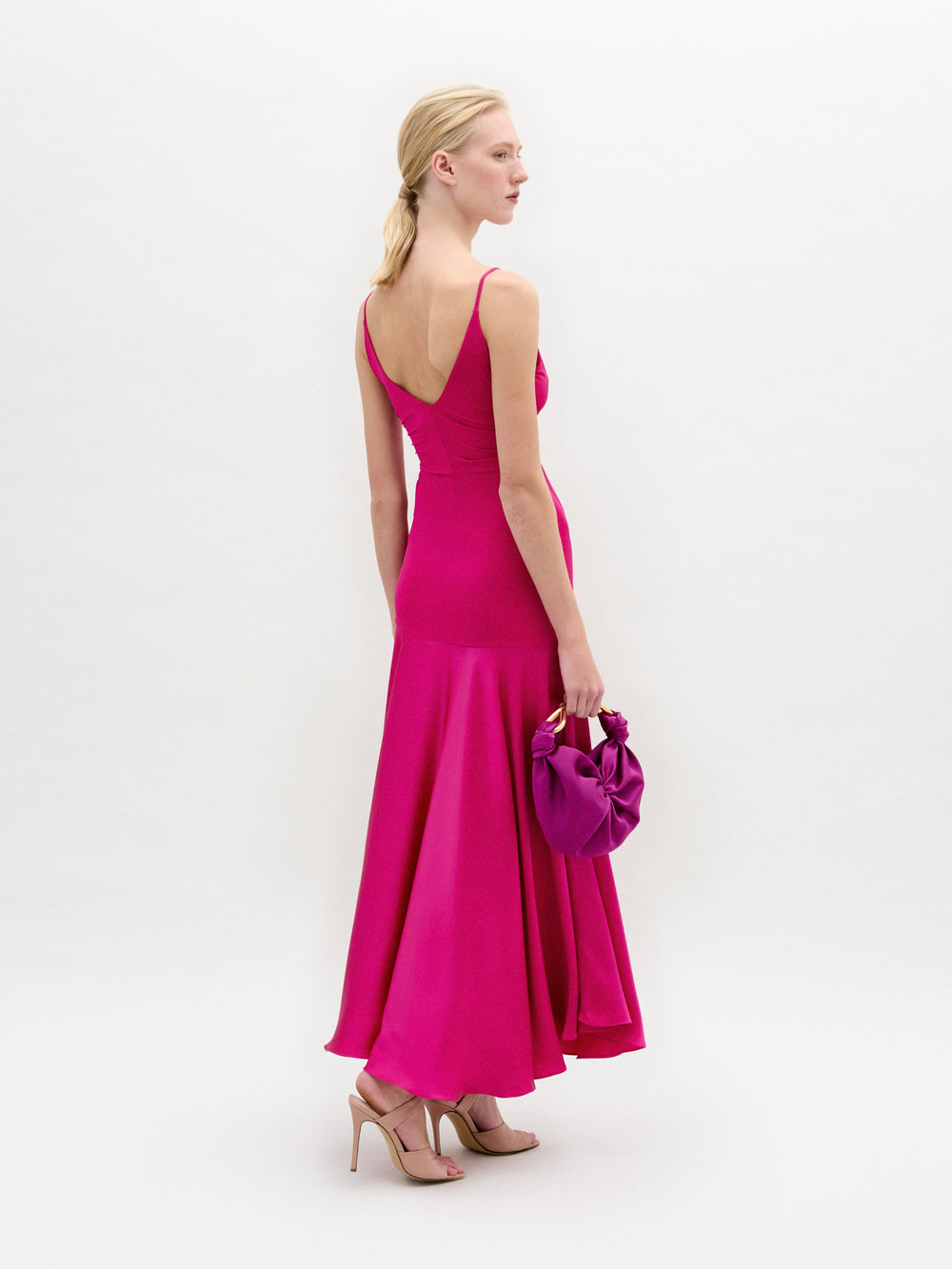 Una mujer con el vestido sin mangas Casiana, de color rosa vivo, confeccionado en tejido italiano, y zapatos de tacón nude, de pie sobre un fondo blanco, con un bolso rosa a juego en la mano izquierda y mirando ligeramente hacia el otro lado de la cámara.