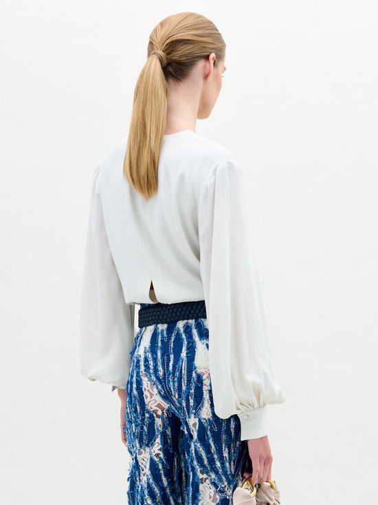 Se ve de espaldas a una persona con el pelo largo de color castaño claro recogido en una cola de caballo. Lleva puesta la Blusa Covilha, una blusa de seda blanca con un elegante escote en forma de ojo de cerradura, y pantalones con estampados azules y blancos. Sujeta un pequeño bolso beige y su conjunto irradia estilo "Hecho en Colombia".