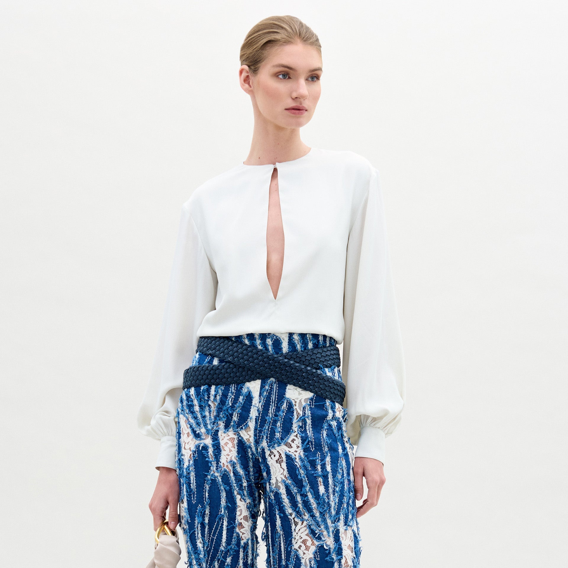Una persona, sobre un fondo liso, lleva la blusa Covilha, un top blanco de seda de manga larga con abertura frontal, combinado con pantalones azules y blancos estampados, y un pequeño bolso en la mano.