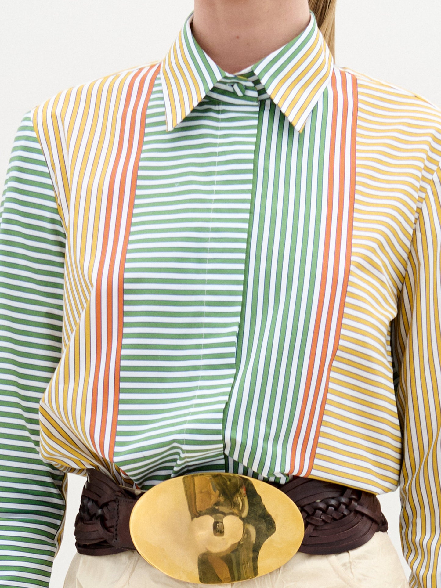 Alguien con una Camisa Cubillos confeccionada en 100% algodón, que muestra rayas verticales en naranja, verde y amarillo. La camisa incluye un cuello y está decorada con un cinturón ancho trenzado de color marrón que presenta una gran hebilla ovalada dorada.