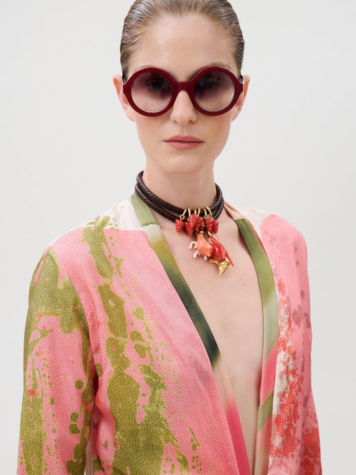 A person wears the Cecelia Tunic in pink and green silk, styled with deep red round sunglasses and a brown leather choker featuring pink and orange organic charms.
