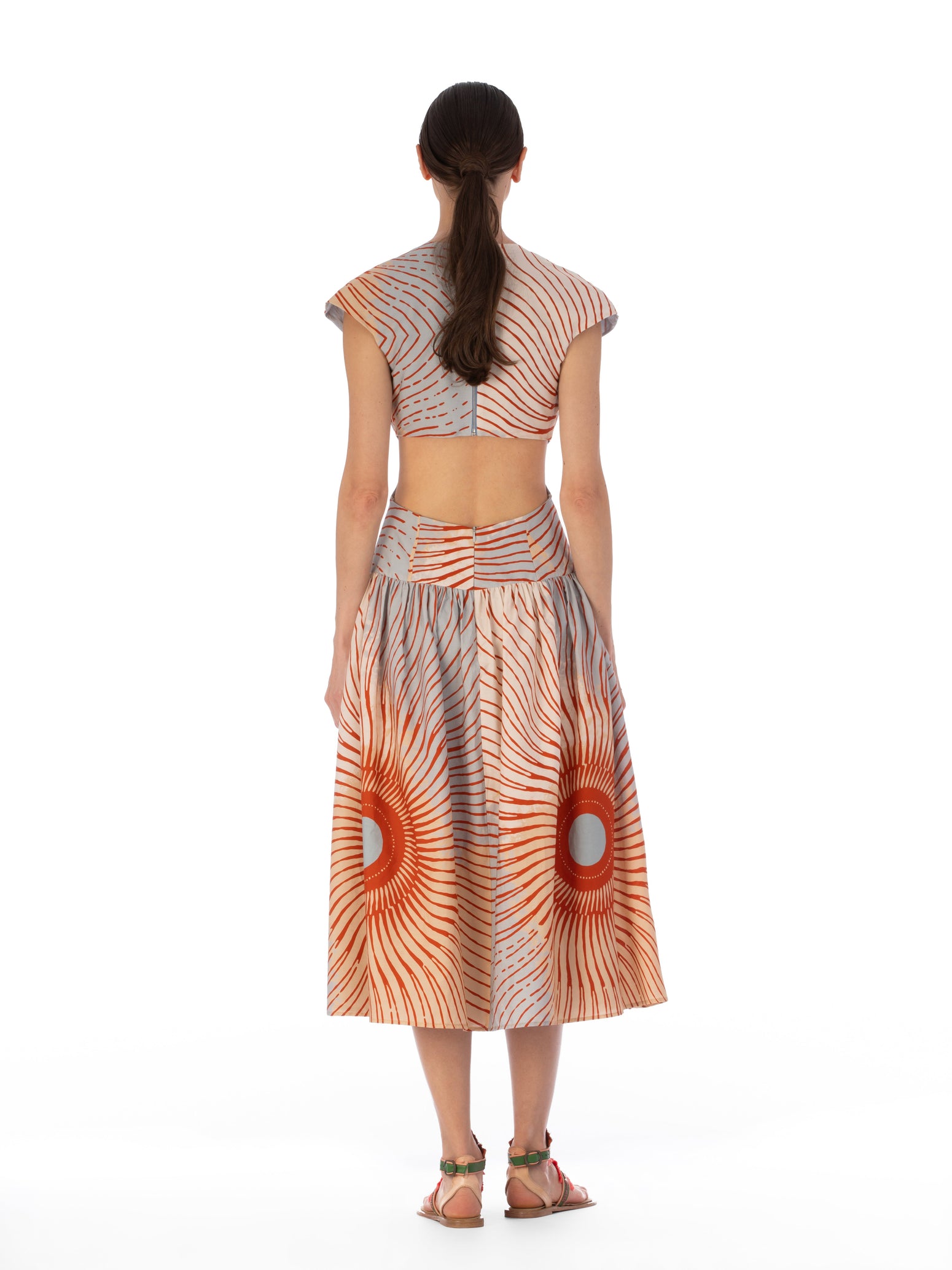 Una mujer de cabello largo y oscuro mira hacia otro lado, vestida con el Vestido Cecile, que presenta un estampado de rayos de sol en color naranja. Lleva un top corto con mangas cortas y una falda midi que recuerda a un estampado multicolor, combinada con sandalias, sobre un fondo blanco liso.