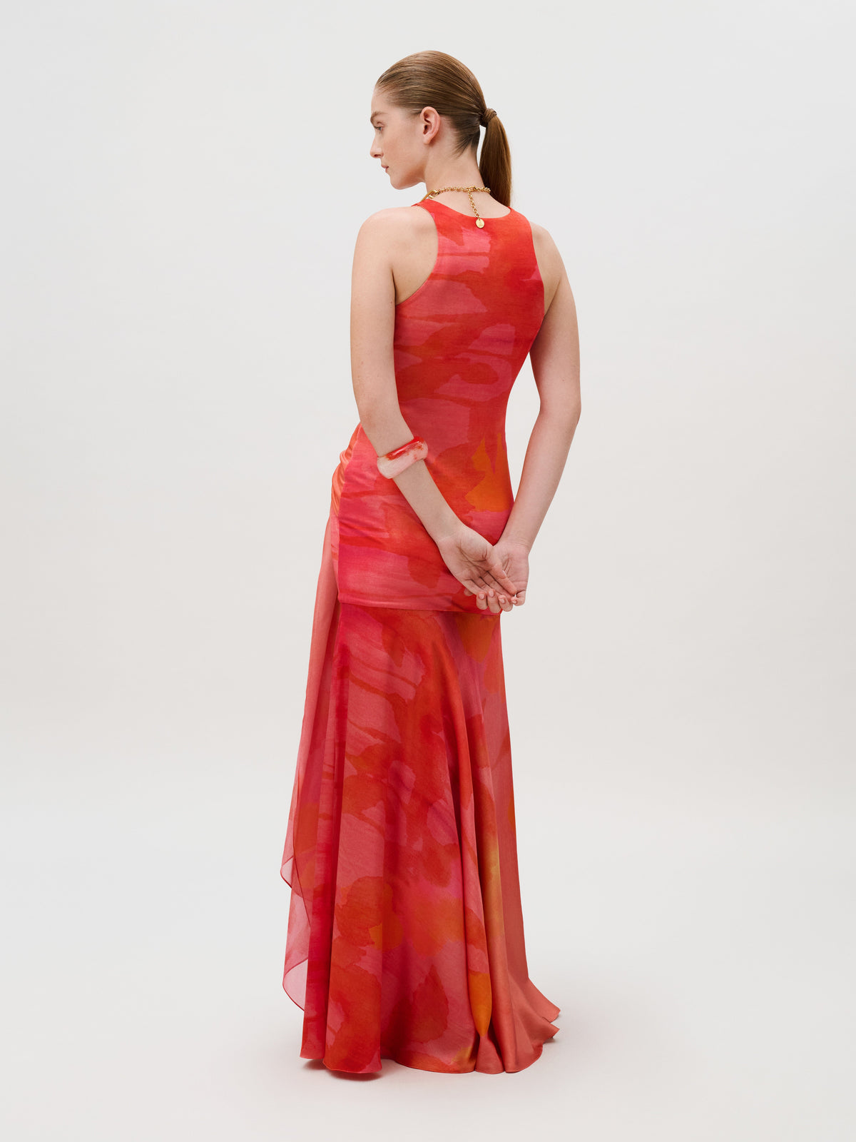 The Clarice Dress is a floor-length, slim-fit gown in a red and orange abstract silk viscose blend, featuring a sleeveless silhouette and a draped side panel.