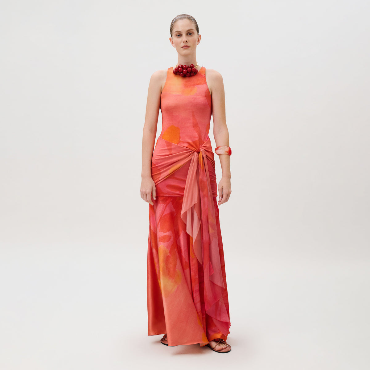 A model wears the Clarice Dress, a floor-length sleeveless silk viscose blend with a red and orange floral print, a knotted waist tie, and a matching red statement necklace.
