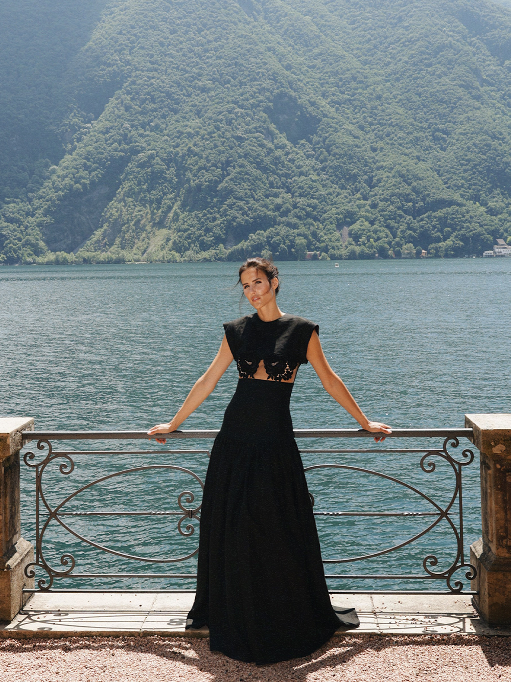 Una mujer con un vestido largo y negro se encuentra de pie frente a una barandilla con un paisaje de fondo que muestra un lago y montañas verdes en un día soleado.
