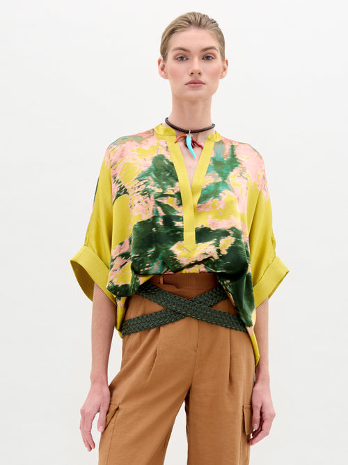 Una persona lleva puesta la Blusa Daia, una blusa con estampado abstracto en amarillo y rosa y mangas anchas en tela italiana de lujo. El atuendo se combina con pantalones color canela, una gargantilla marrón y detalles verdes de un cinturón trenzado sobre un fondo claro y sencillo.