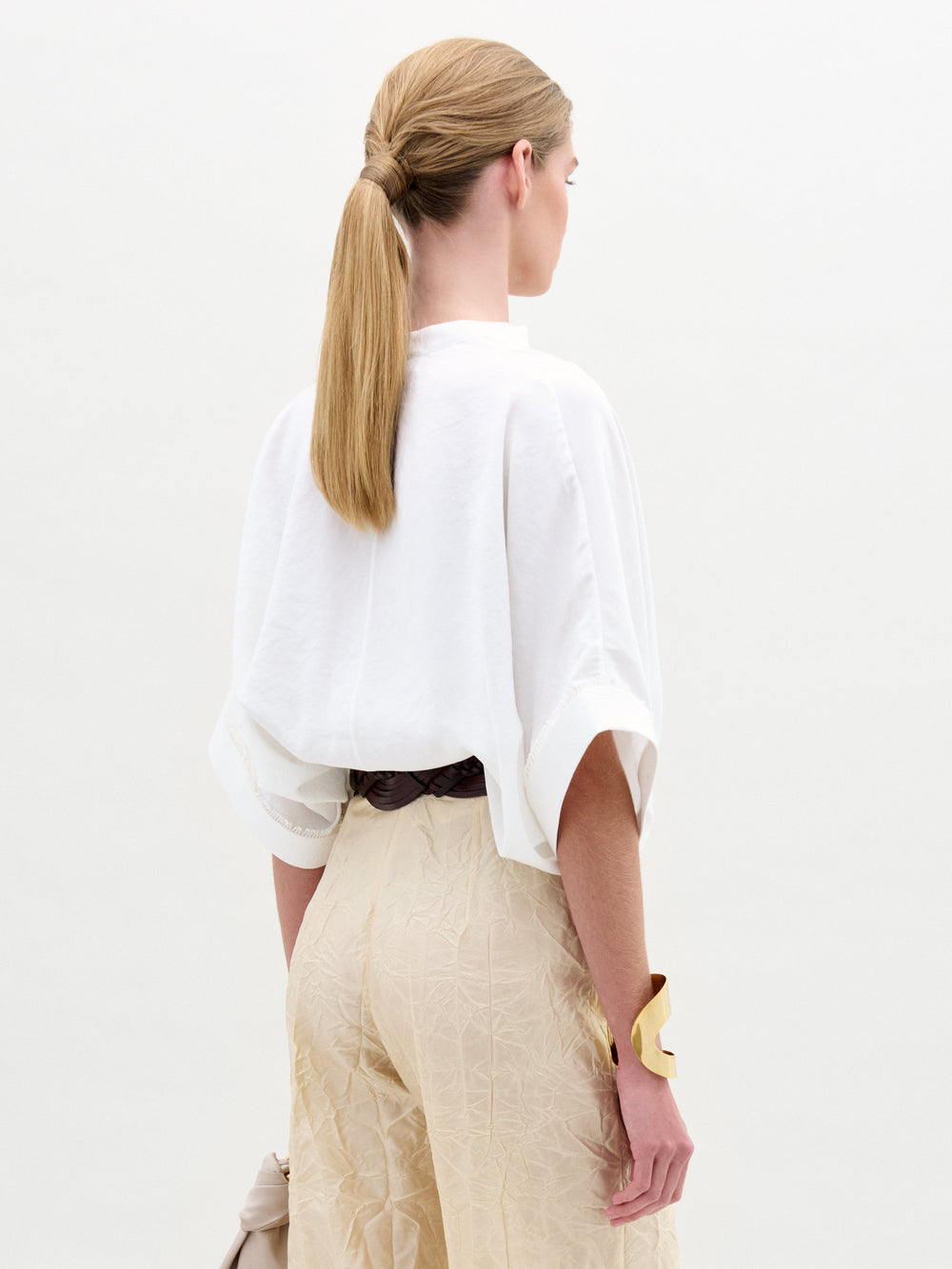 Una persona con el pelo largo y rubio, atado en una coleta baja, está de espaldas. Lleva una blusa Daia con mangas arremangadas y pantalones beige. Este look suelto de top se combina elegantemente con una pulsera dorada y un bolso de color claro sobre un fondo blanco liso.