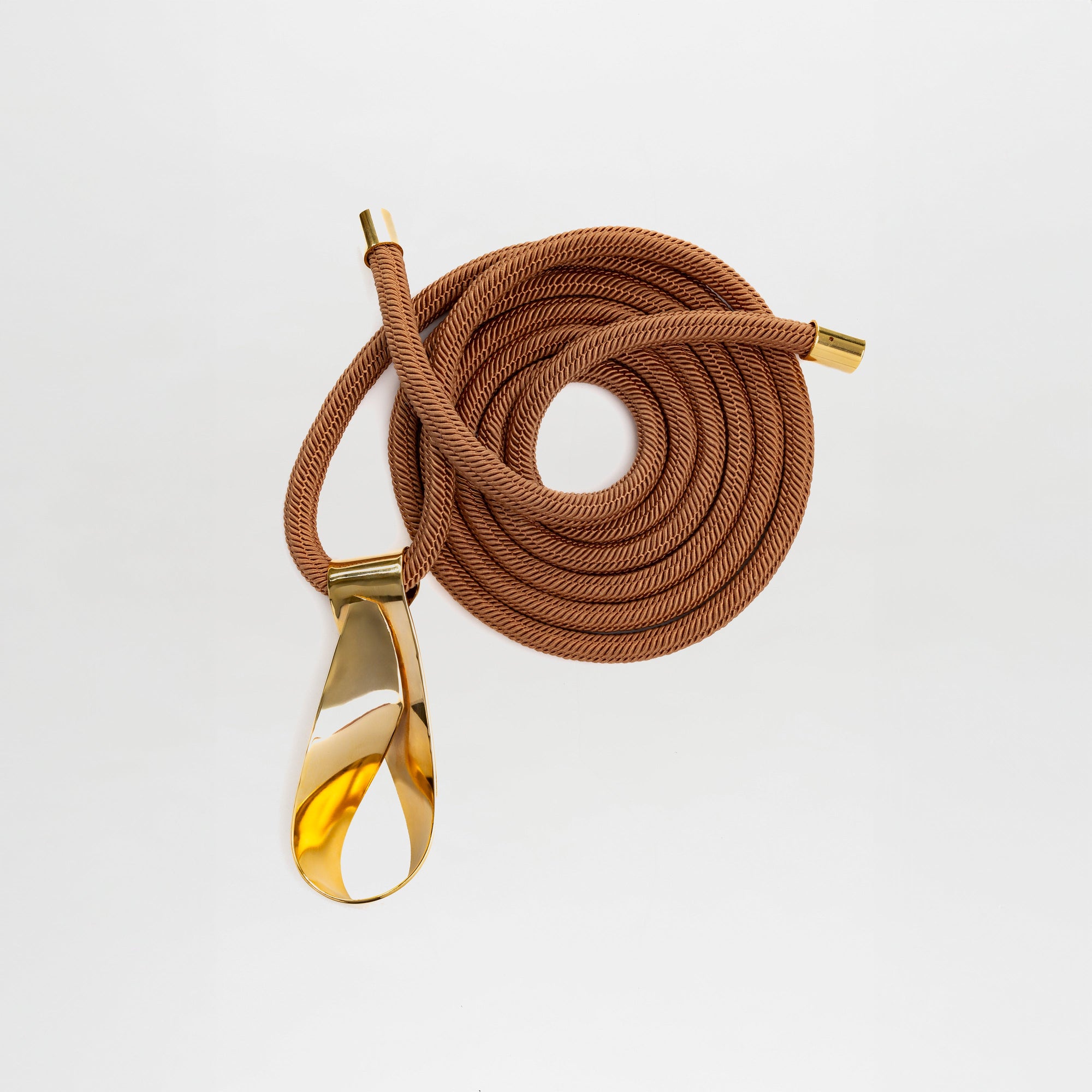 El Cinturón Daiana, fabricado en Colombia, presenta un cordón marrón enrollado con puntas metálicas doradas y una gran hebilla dorada pulida en forma de lágrima, desplegada sobre un fondo blanco liso.