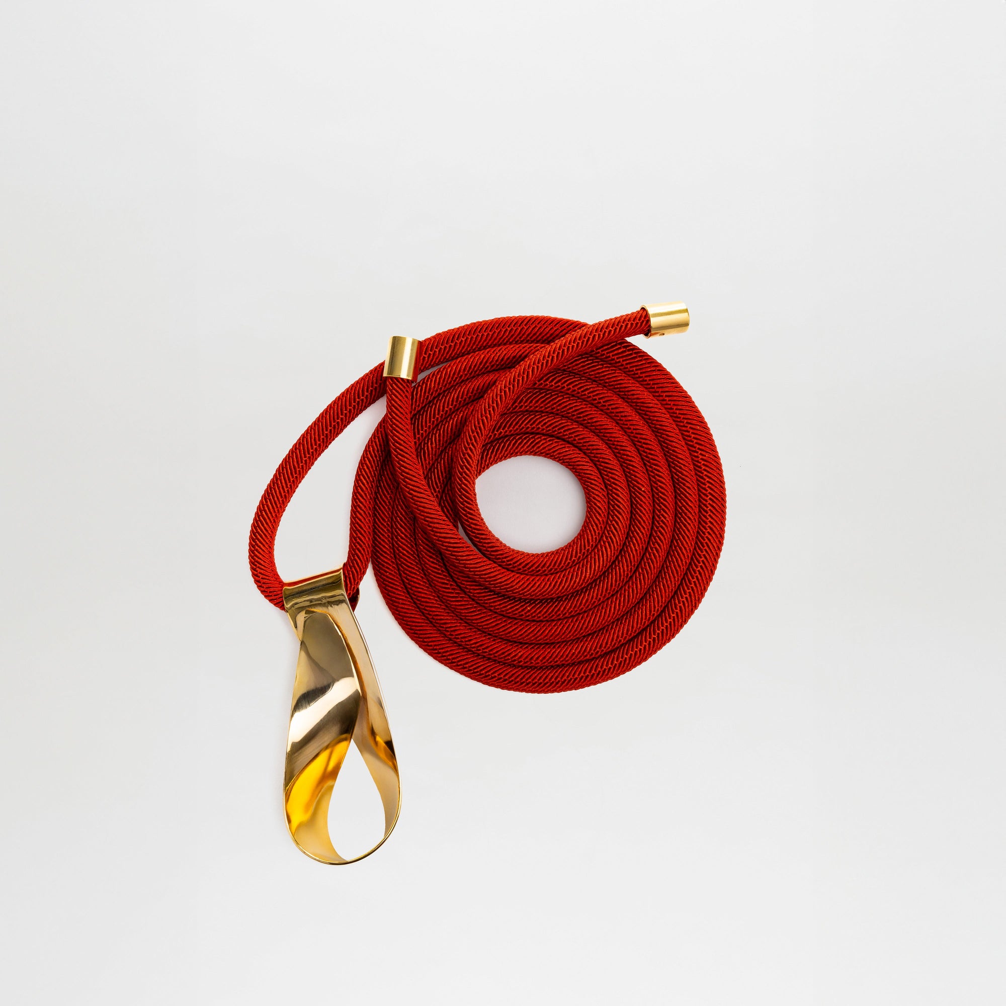 El Cinturón Daiana, un cinturón de cordón rojo enrollado con extremos de metal dorado y un gran lazo dorado, se muestra sobre un fondo blanco liso.