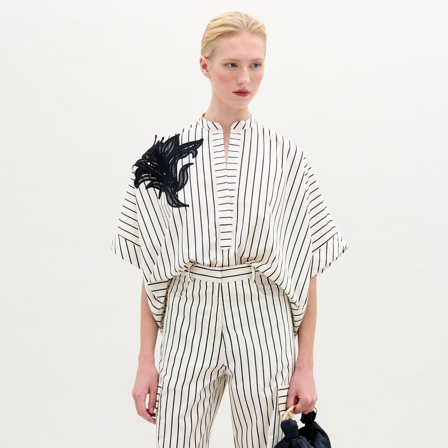 Una persona luce la blusa Daia, una blusa blanca de manga corta 100% algodón con rayas y un detalle decorativo negro en el hombro, combinada con un pantalón a rayas a juego sobre un fondo blanco liso.