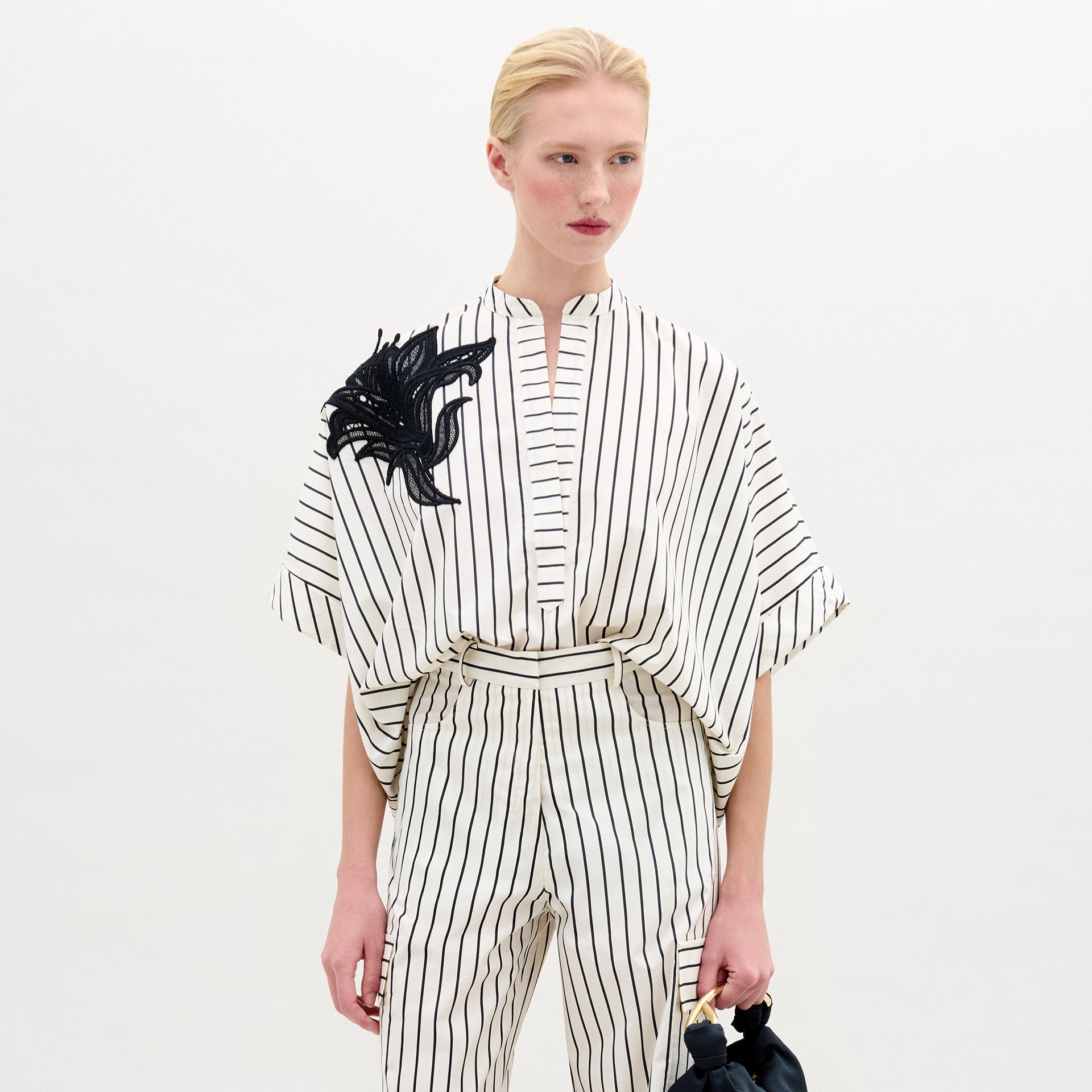 Una persona luce la blusa Daia, una blusa blanca de manga corta 100% algodón con rayas y un detalle decorativo negro en el hombro, combinada con un pantalón a rayas a juego sobre un fondo blanco liso.