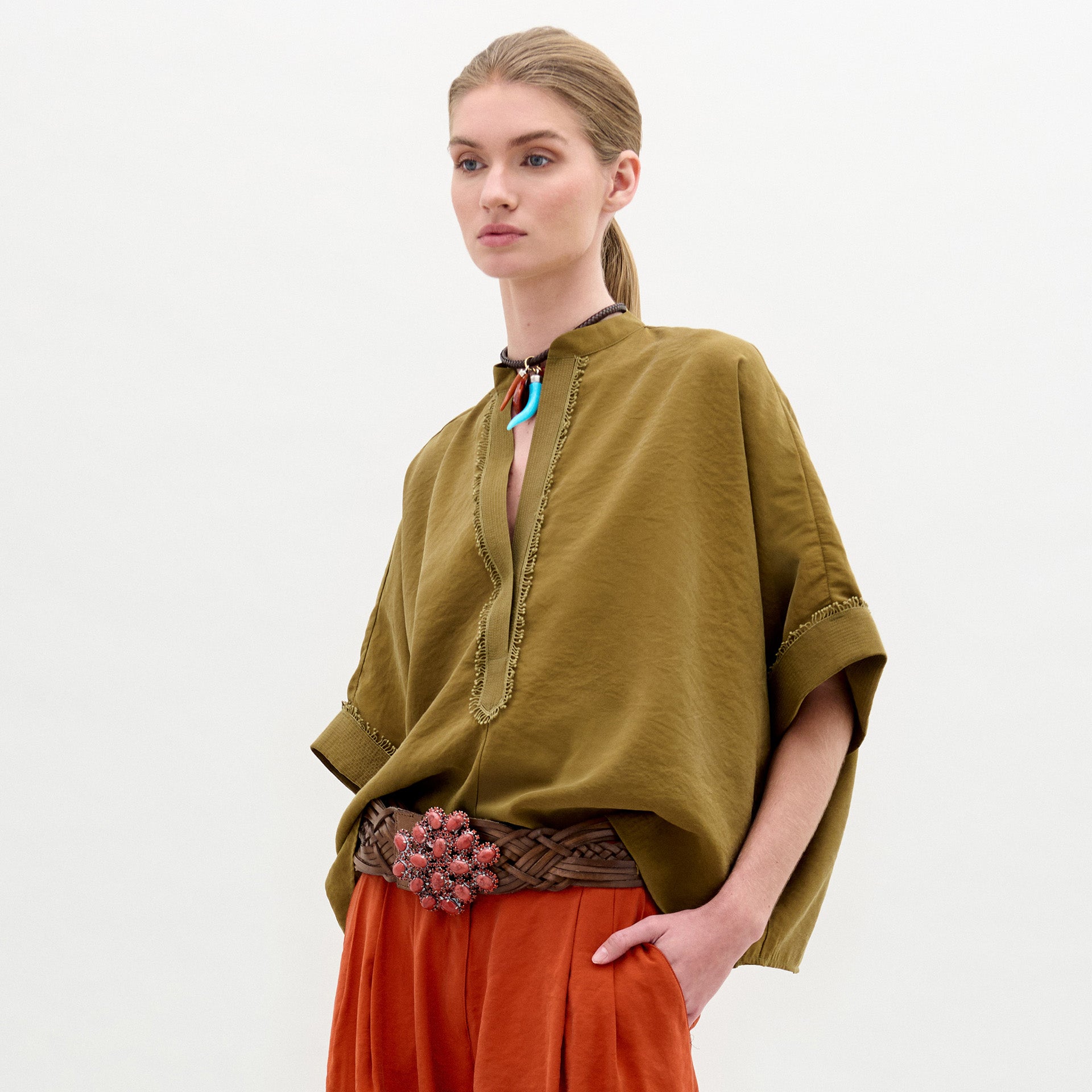 Una persona de pie sobre un fondo blanco liso lleva una blusa Daia de corte amplio en verde oliva con mangas enrolladas, pantalones naranja rojizo, un cinturón marrón tejido con una hebilla floral y un collar con acentos azules y rojos.