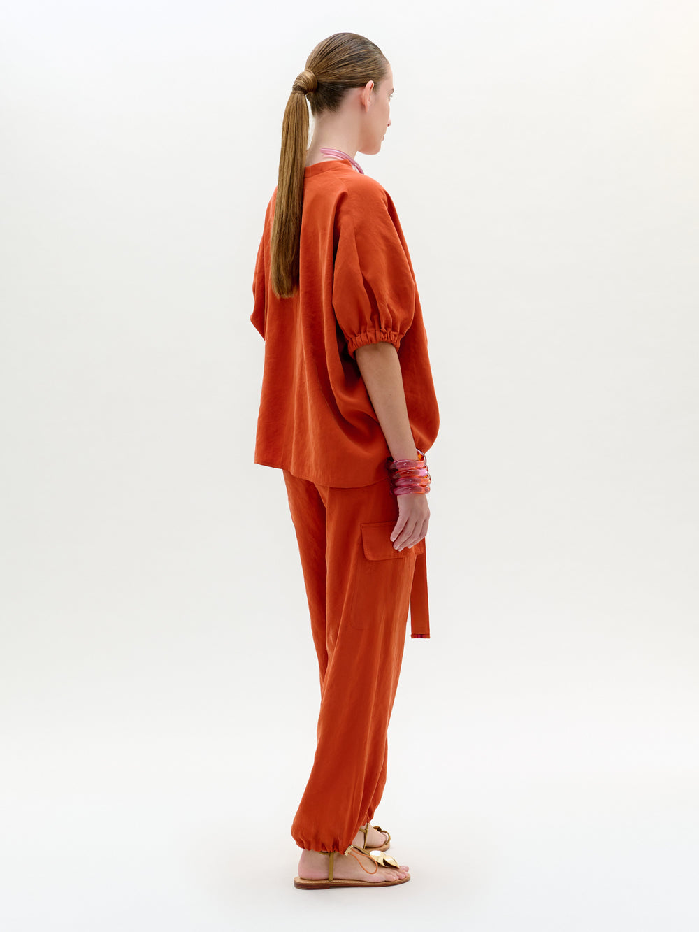 Una persona aparece de perfil sobre un fondo liso, vestida con un top naranja holgado de mangas abullonadas y pantalones Darnell a juego. Lleva el pelo largo recogido y accesorios como sandalias y pulseras.