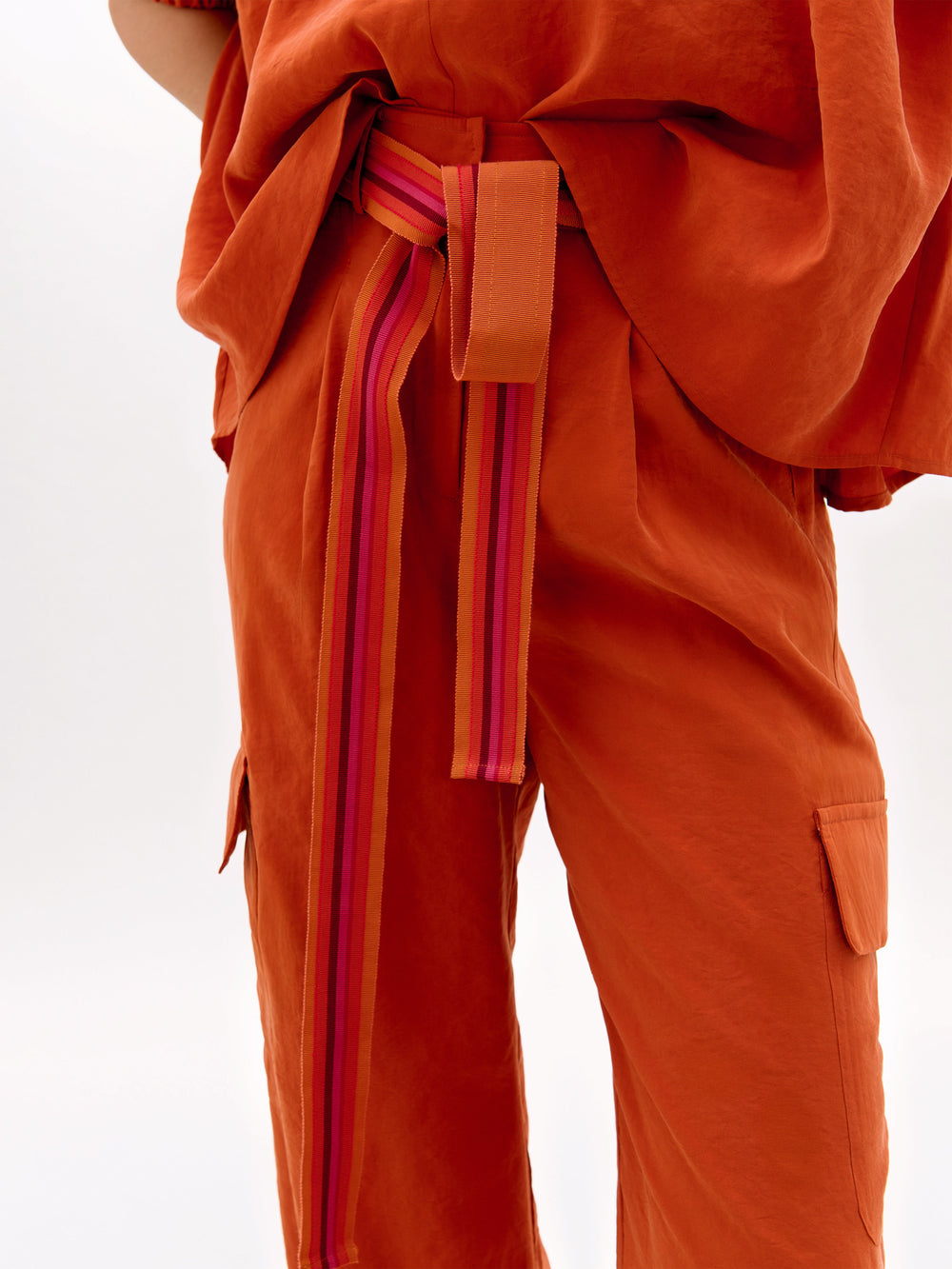Una persona, sobre un fondo liso, lleva un pantalón Darnell naranja holgado y un top a juego. El conjunto está adornado con un cinturón de rayas rosas y rojas. Sólo se muestran el torso y la parte superior de las piernas.