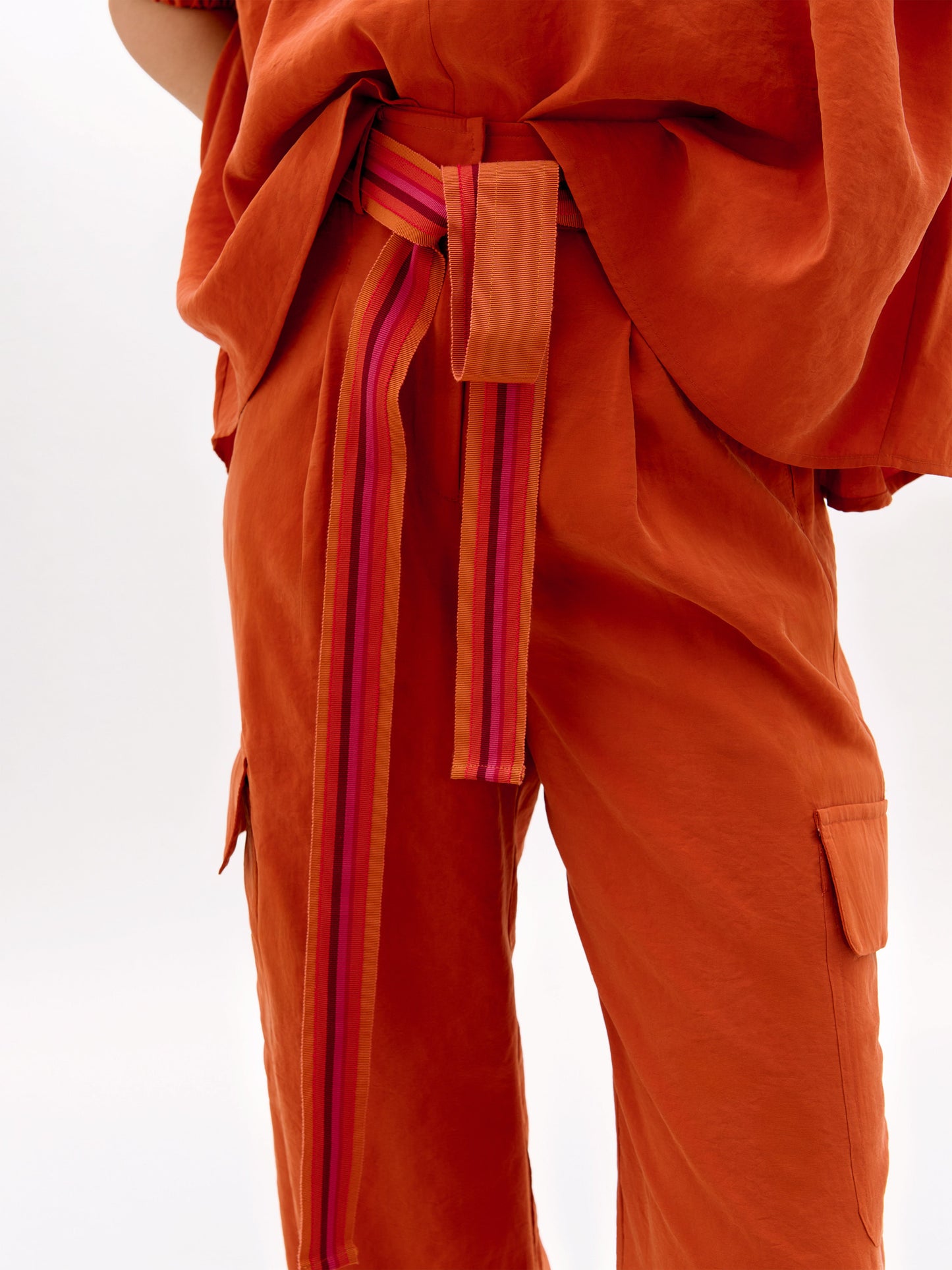 Una persona, sobre un fondo liso, lleva un pantalón Darnell naranja holgado y un top a juego. El conjunto está adornado con un cinturón de rayas rosas y rojas. Sólo se muestran el torso y la parte superior de las piernas.