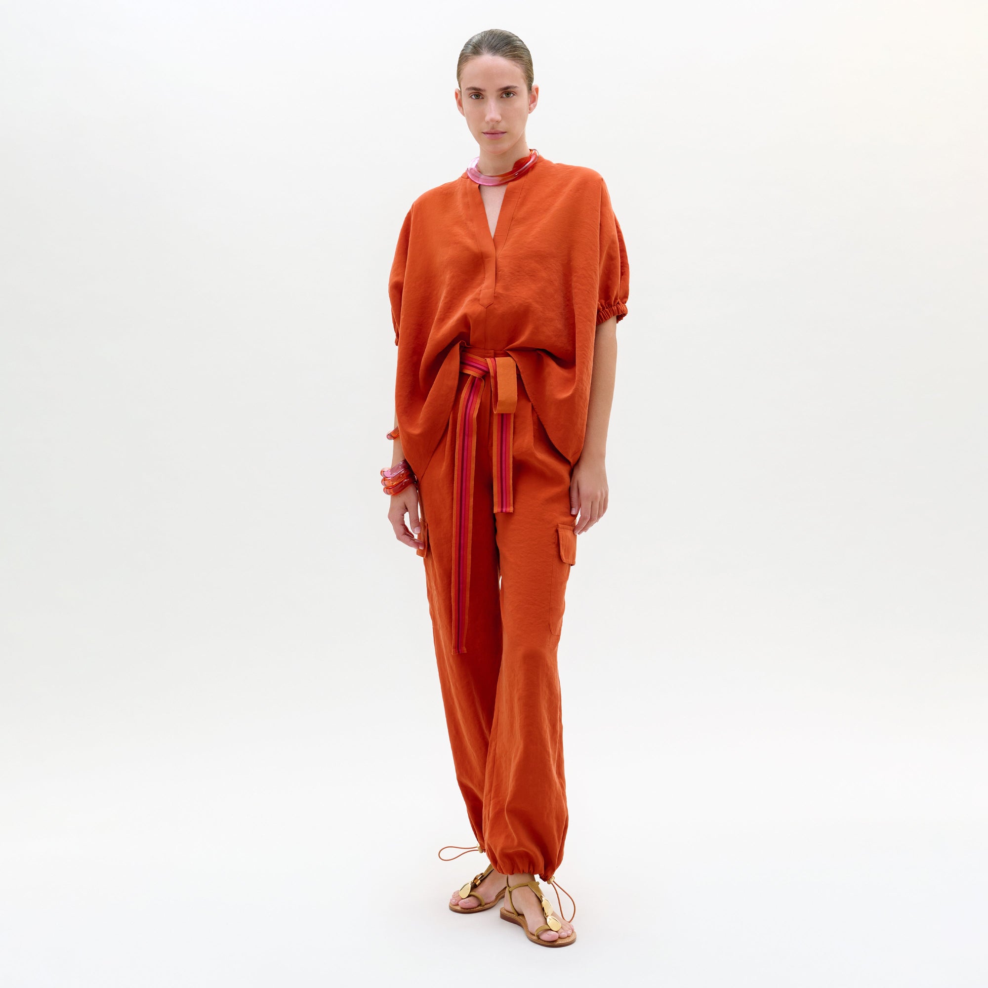 Una persona de pie sobre un fondo blanco liso lleva el Darnell Pant, un pantalón cargo naranja de tela italiana de corte holgado con lazos en los tobillos y cintura anudada, combinado con sandalias planas. Llevan una expresión neutra y joyas mínimas.
