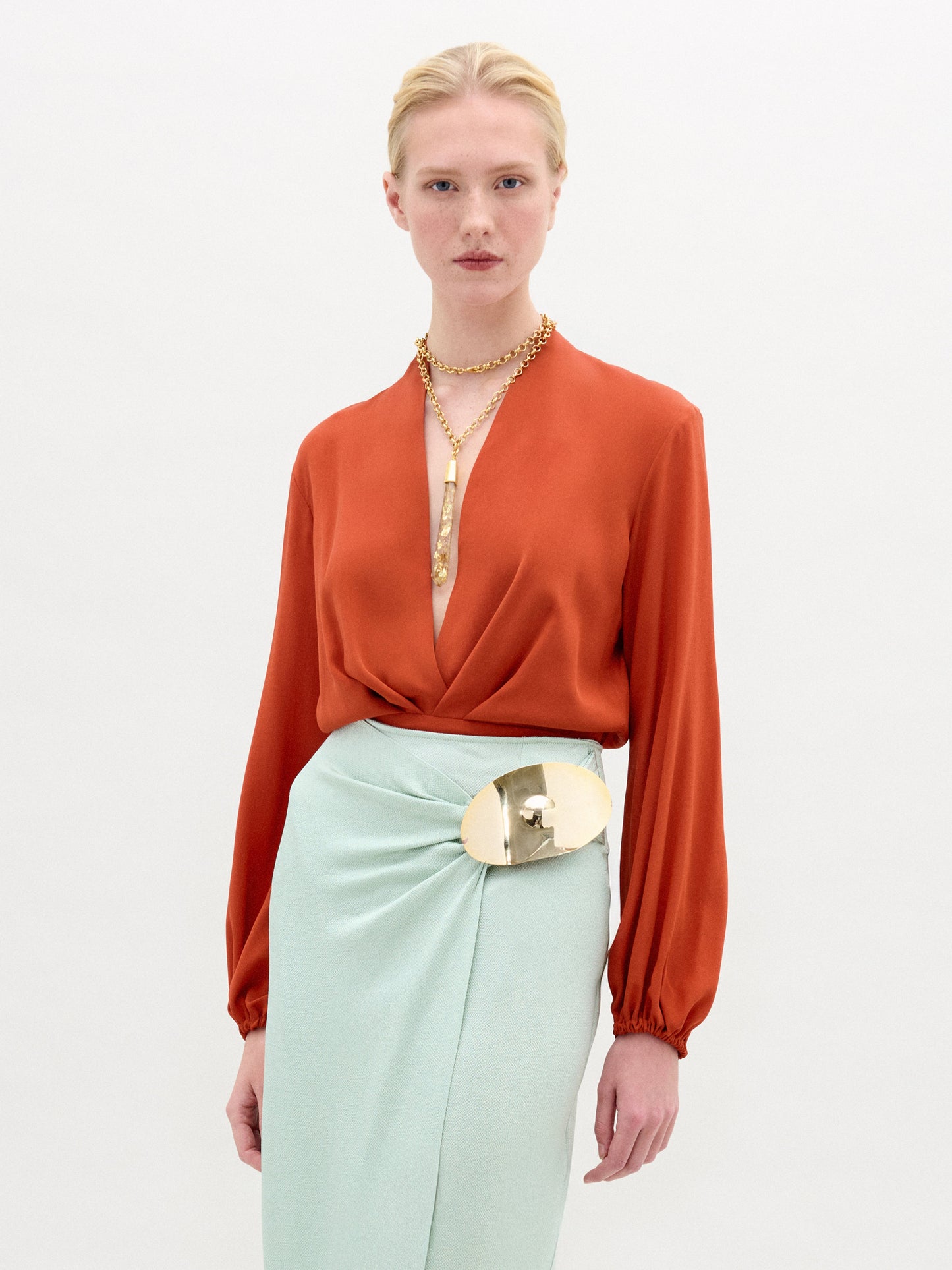 Una persona de pelo rubio lleva la blusa Delfina de seda naranja intenso con una falda verde menta claro y una gran hebilla de cinturón dorada. Los collares dorados en capas acentúan el look sobre un fondo blanco liso. Hecho en Colombia.