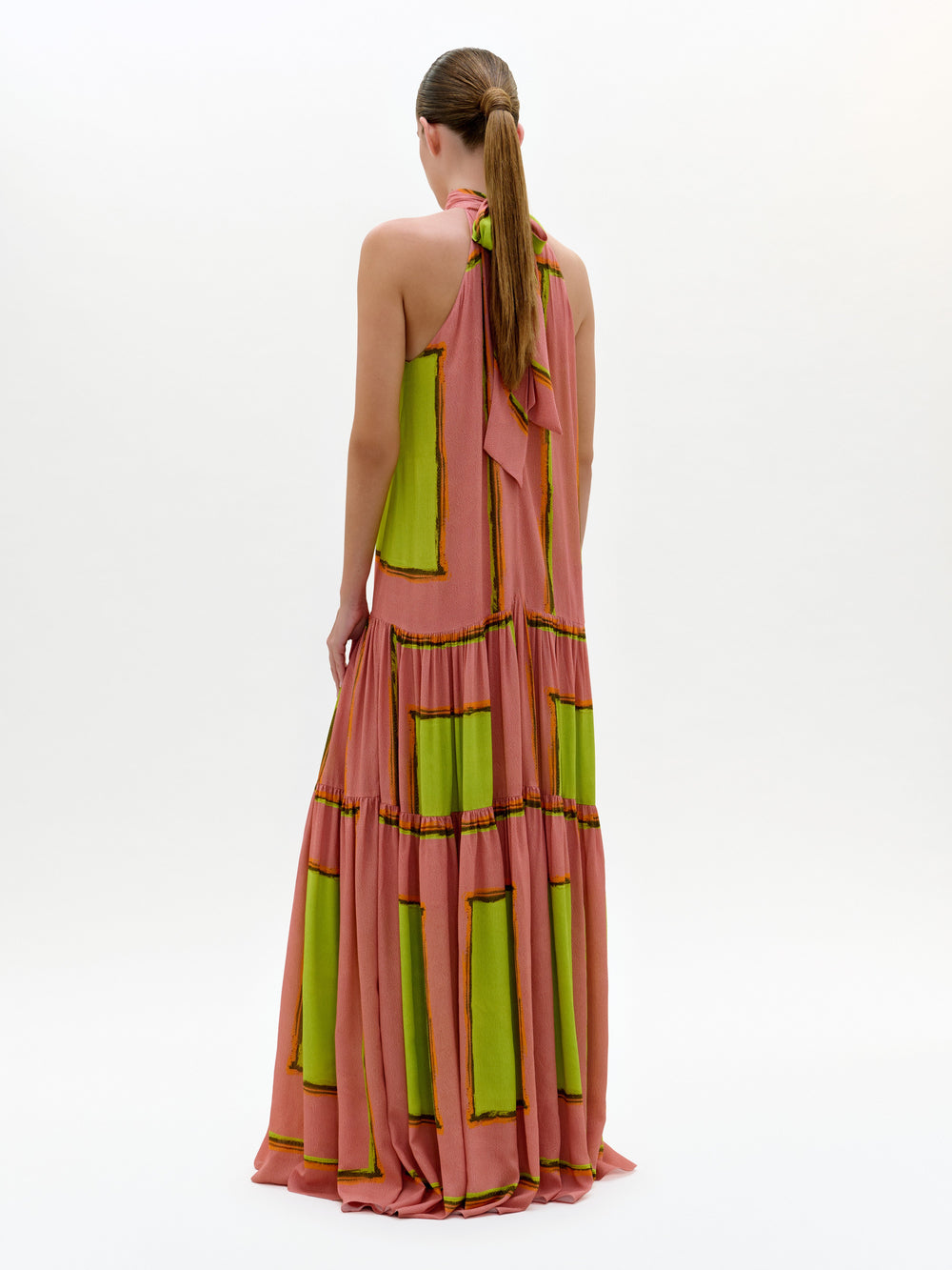 Una persona de espaldas lleva el Dorelia Dress, un vestido maxi rosa con patrones verdes rectangulares y escote halter. Tiene el cabello castaño en coleta baja y posa frente a un fondo blanco liso. Hecho en Colombia.
