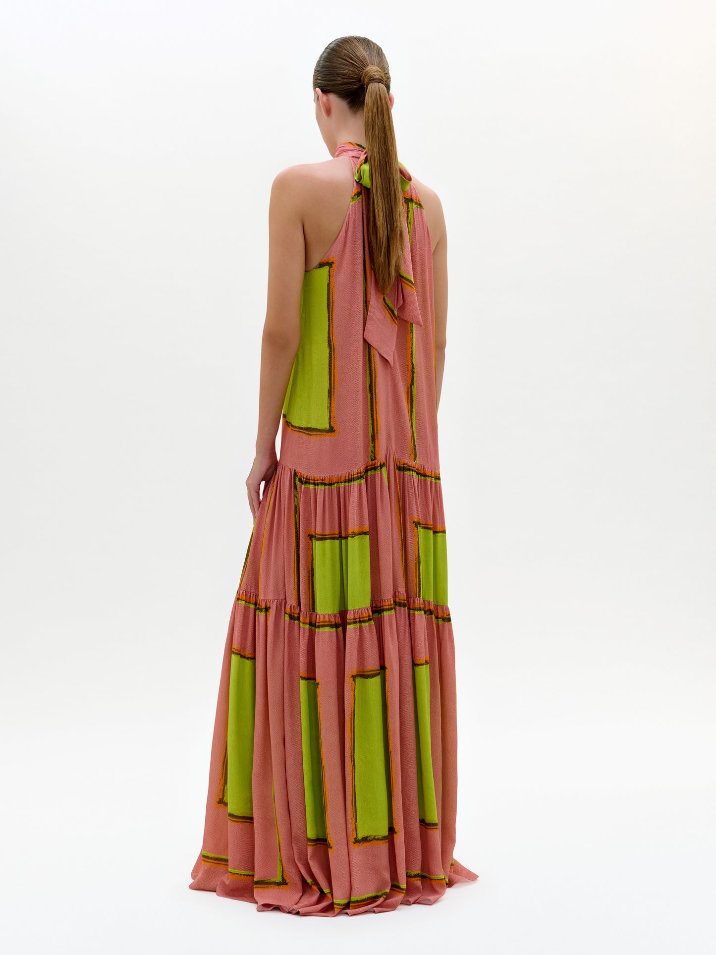 Una persona de espaldas lleva el Dorelia Dress, un vestido maxi rosa con patrones verdes rectangulares y escote halter. Tiene el cabello castaño en coleta baja y posa frente a un fondo blanco liso. Hecho en Colombia.
